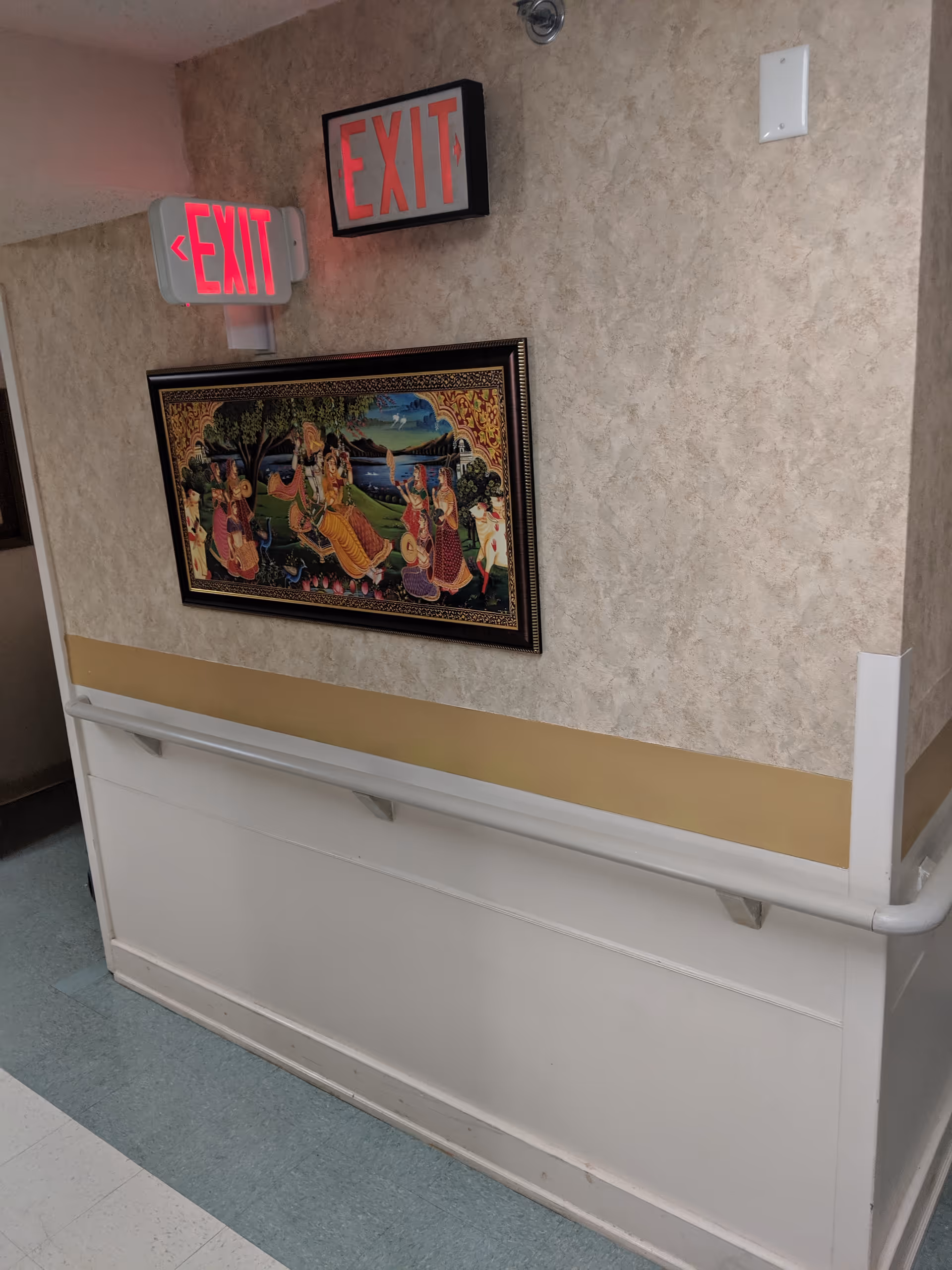 Interior hallway with exit signs, framed artwork, beige walls, and a white railing.