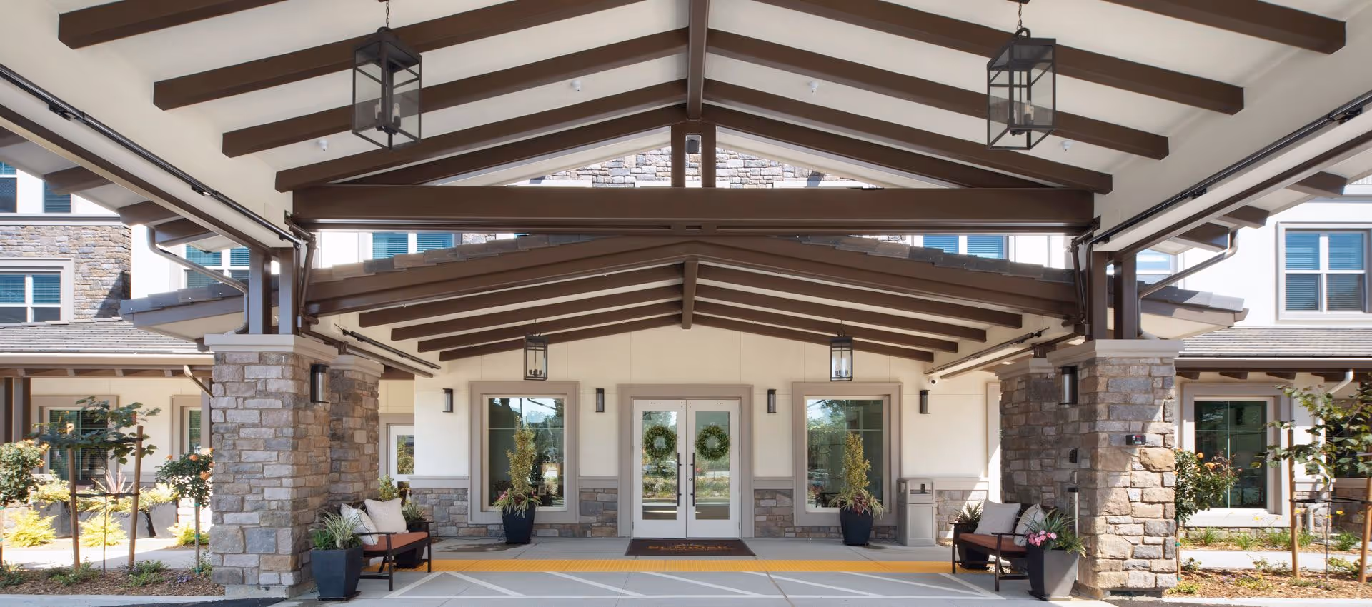 Covered entrance/porte-cochere with exposed wooden beams, stone columns, benches and potted plants at the front of a facility.