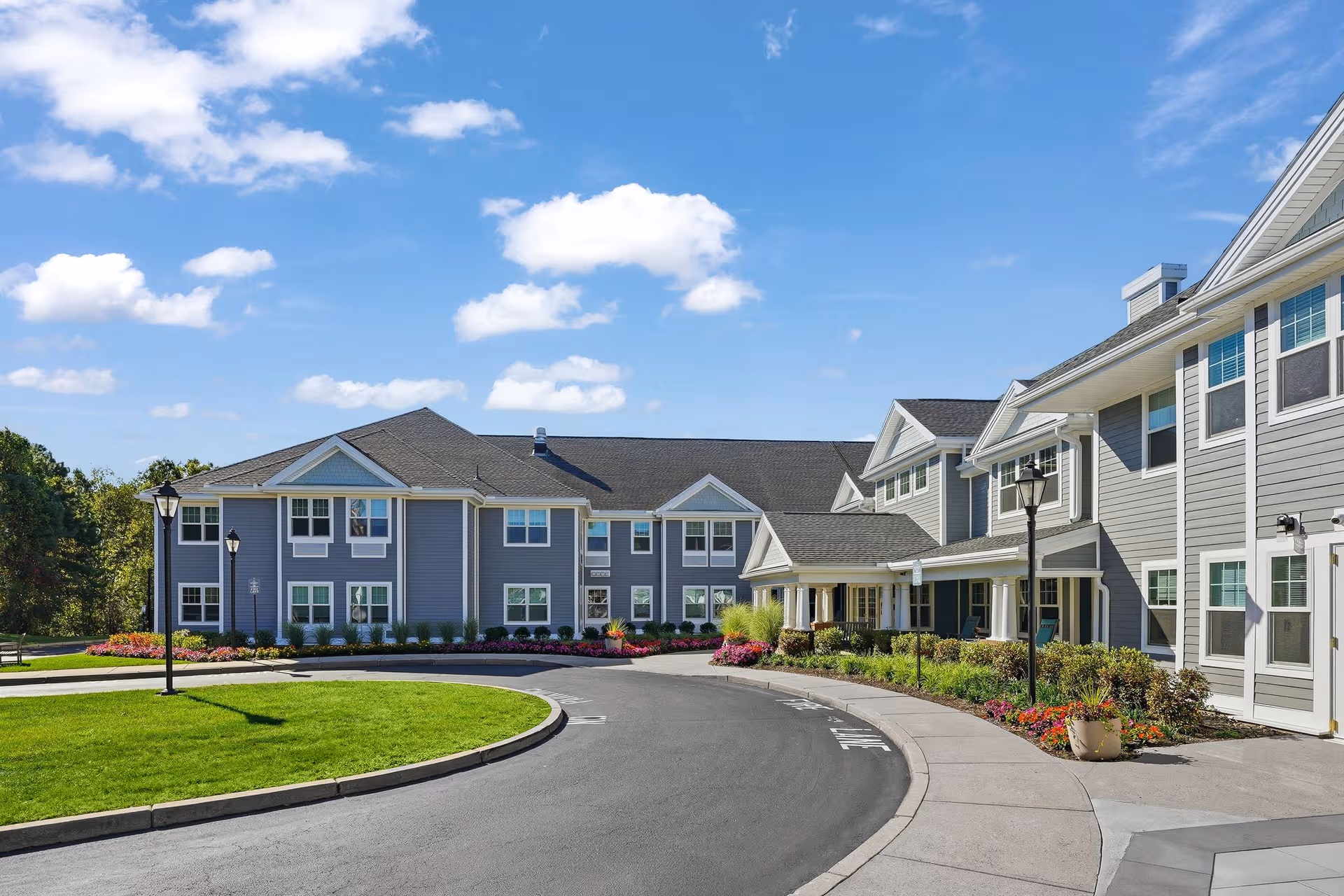 Blue-gray senior living building with curved driveway, manicured landscaping, and lampposts beneath a sunny sky.