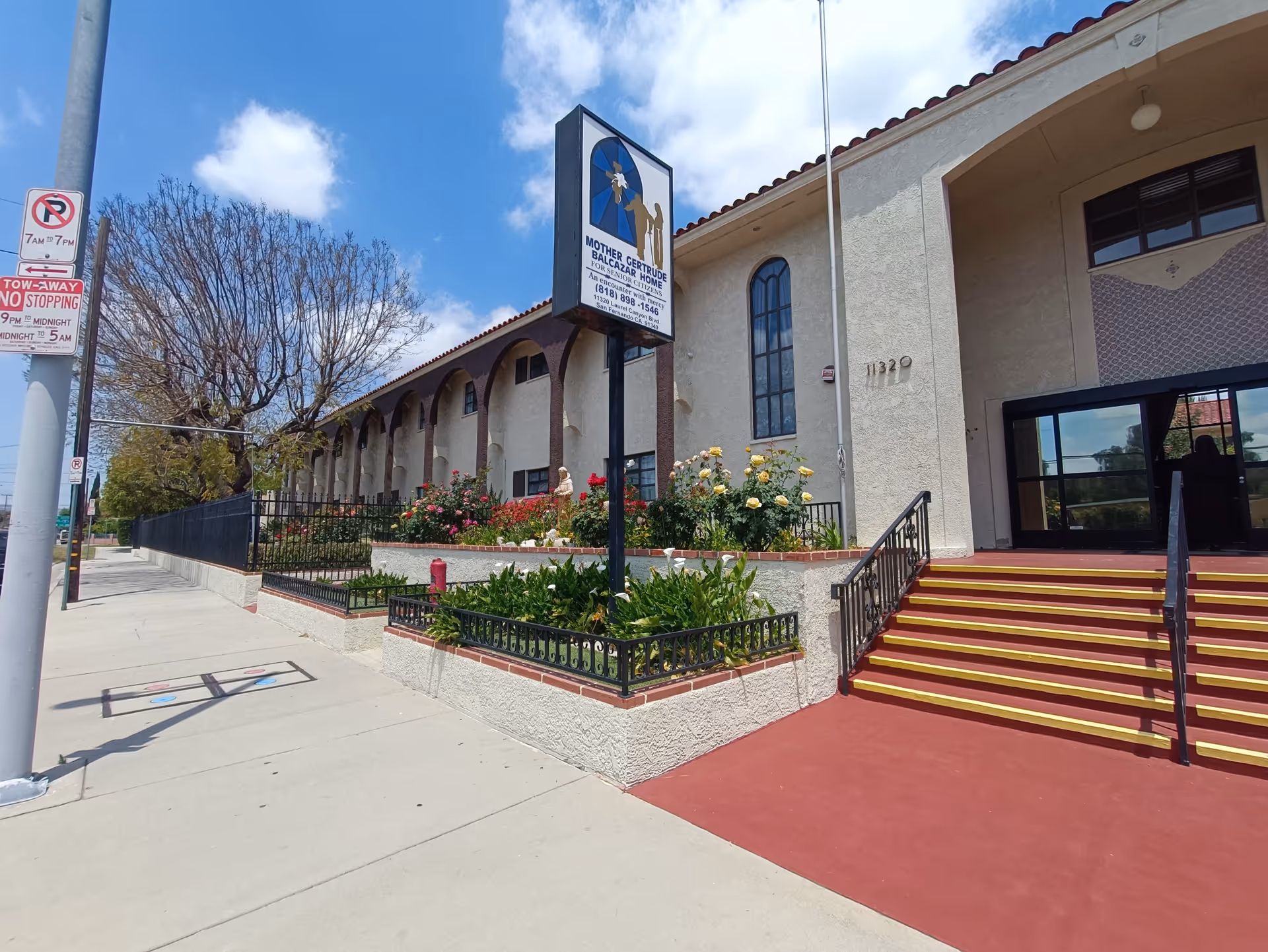 Exterior view of Mother Gertrude Balcazar Home building with a sidewalk, stairs with yellow safety stripes, a garden with flowers, and a sign displaying the facility name and contact information under a partly cloudy sky.