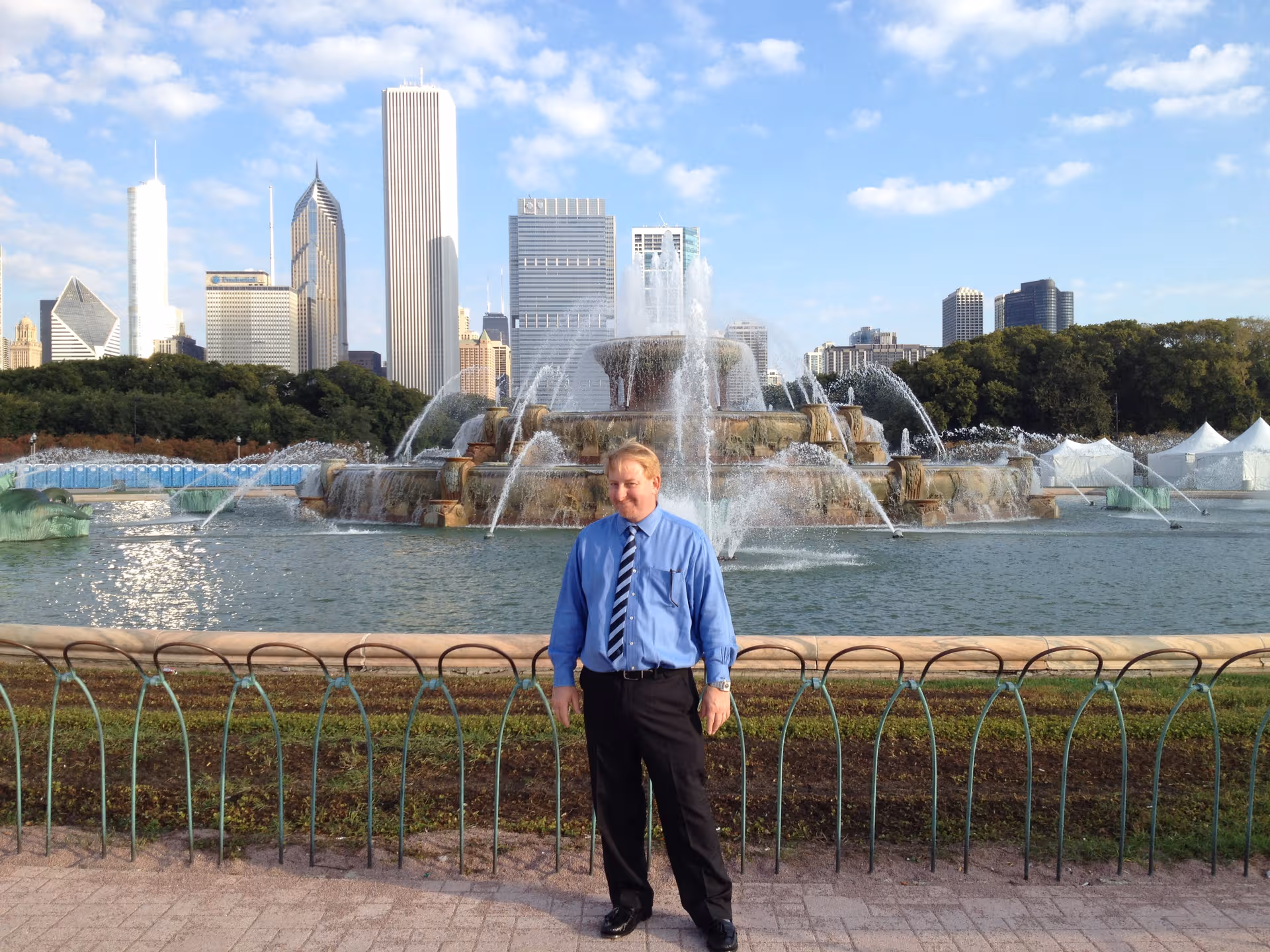 A man in a blue shirt stands beside a fountain with a city skyline behind.