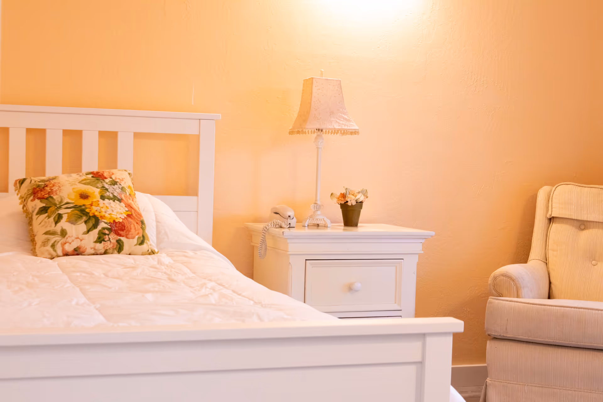 White bed, nightstand, lamp, and beige chair in a peach-colored bedroom.