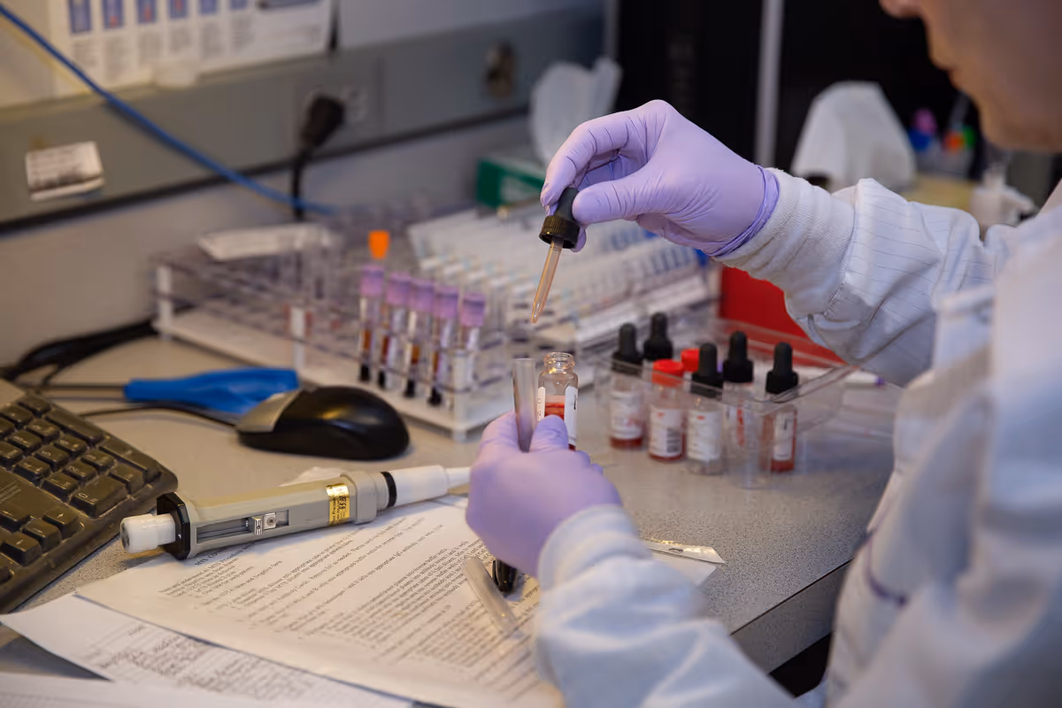 A lab technician wearing gloves handles a vial with a pipette at a bench.