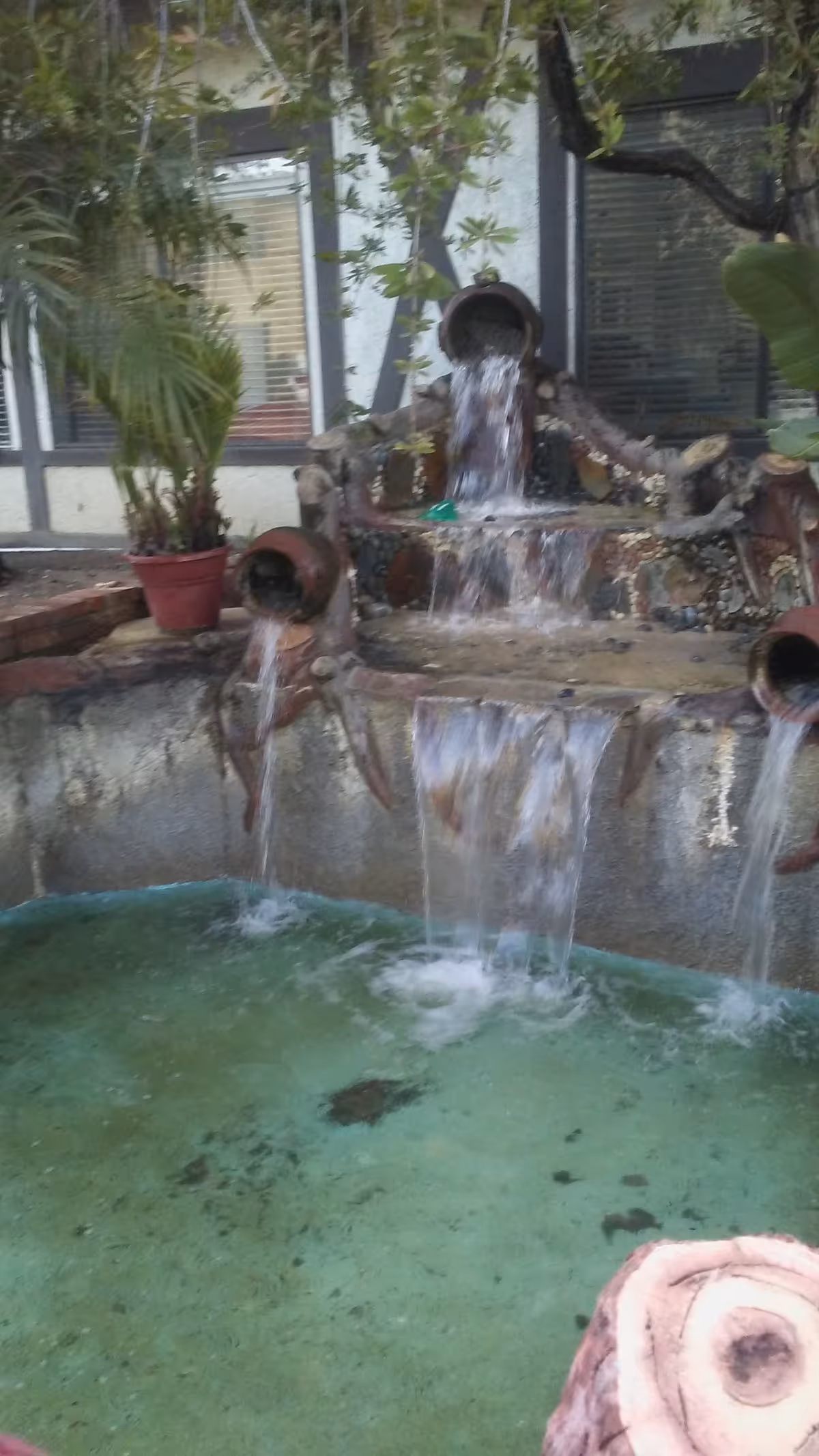 Outdoor courtyard fountain with cascading water, potted plants, and building windows in background.