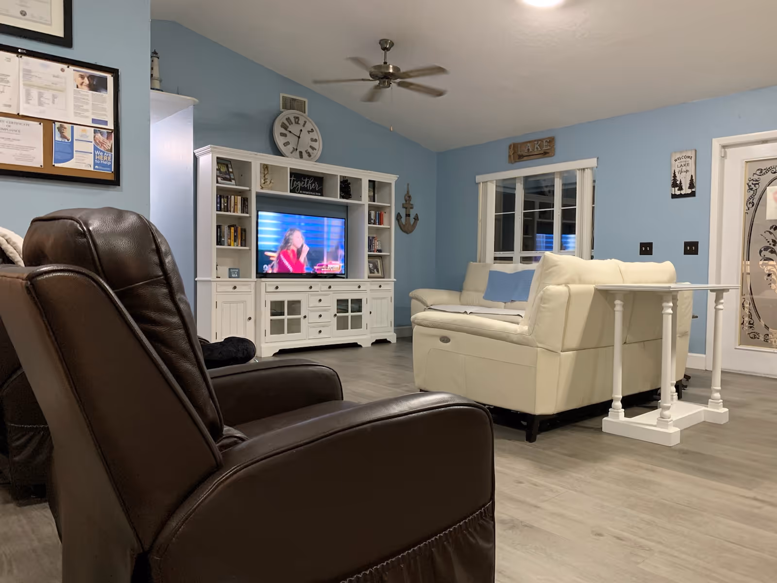A cozy living room with light blue walls, a white entertainment center with a TV showing a woman in a red outfit, a large wall clock above it, and shelves with books and decor. There is a white leather sofa with blue pillows, a small white table behind it, a brown leather recliner in the foreground, and a ceiling fan above. The room has wood flooring and a window with white blinds.