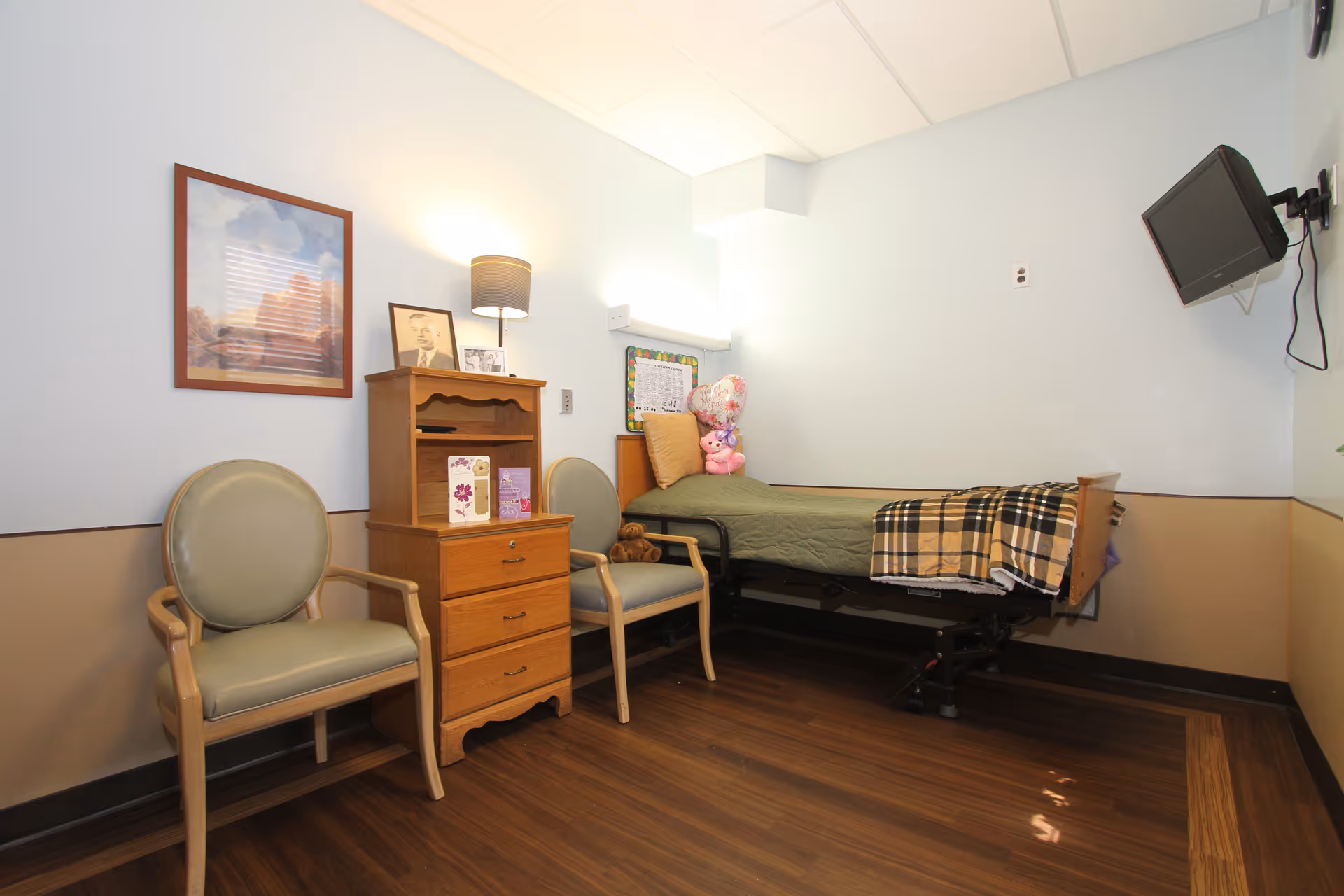A small, neatly arranged room with a single hospital-style bed covered with a green blanket and a plaid throw. Two beige chairs with wooden arms are placed next to a wooden dresser with three drawers, a lamp, and framed pictures on top. A small TV is mounted on the wall opposite the bed. The room has light blue walls with a beige lower half and wood flooring.