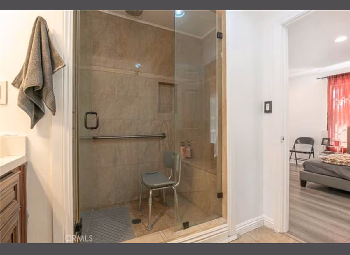 Glass shower enclosure in a bathroom; towel hangs, vanity on the left, bedroom beyond.