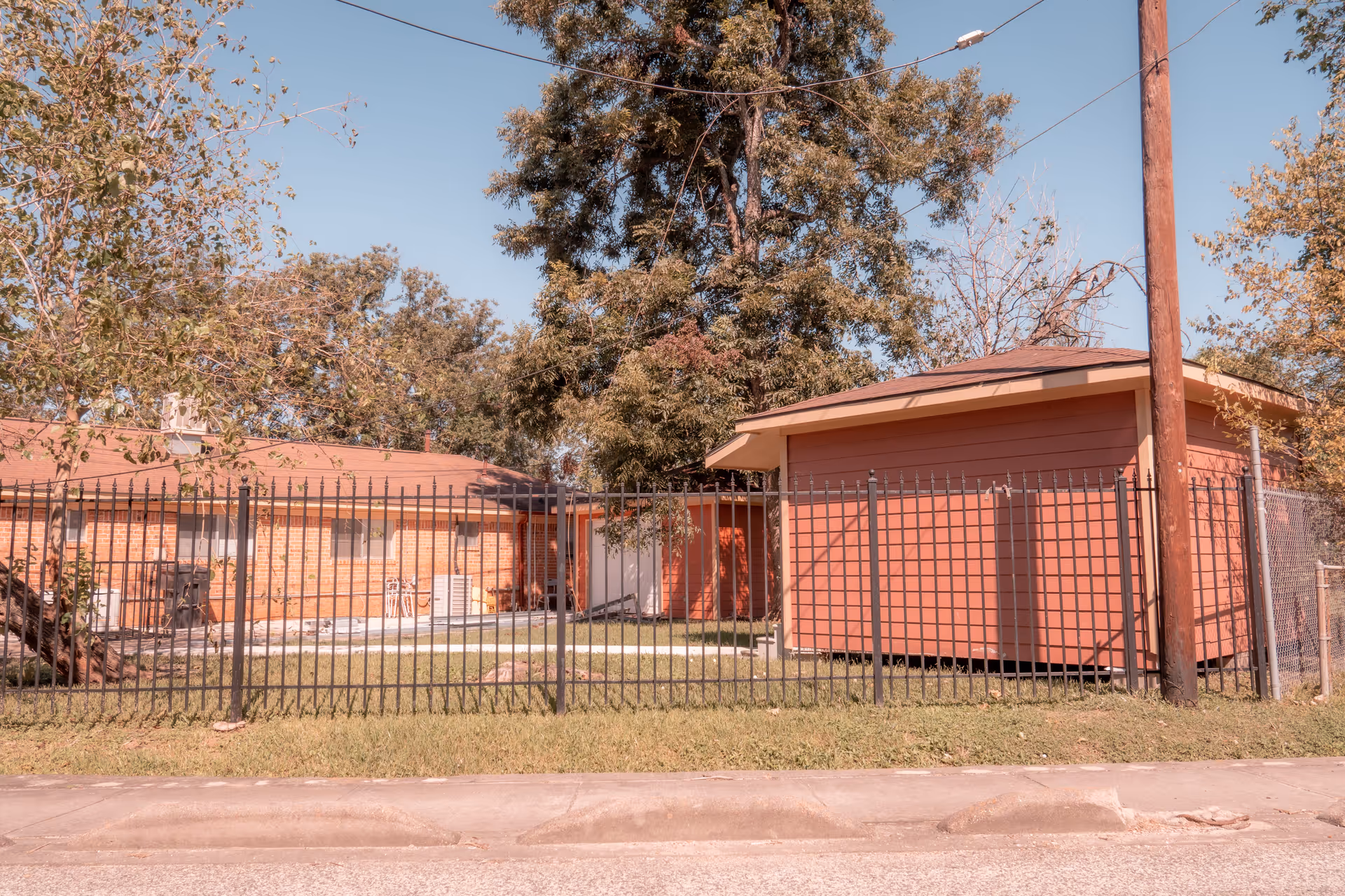 Single-story brick building and a small red outbuilding behind a black metal fence, with trees and a utility pole in front.