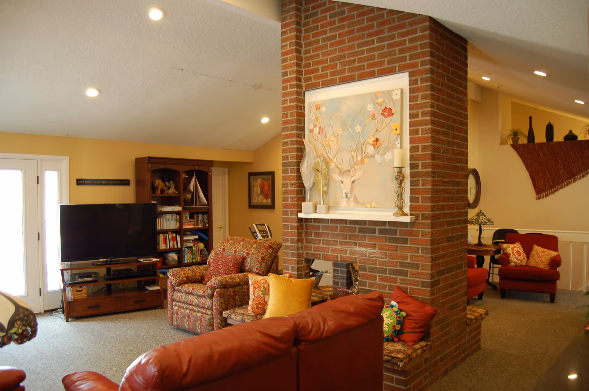 A cozy living room with a brick fireplace in the center. The room features a red leather sofa, a patterned armchair, and a wooden TV stand with a flat-screen television. There is a bookshelf with various items and books, and decorative artwork above the fireplace. The walls are painted a warm yellow, and the room is well-lit with recessed ceiling lights.
