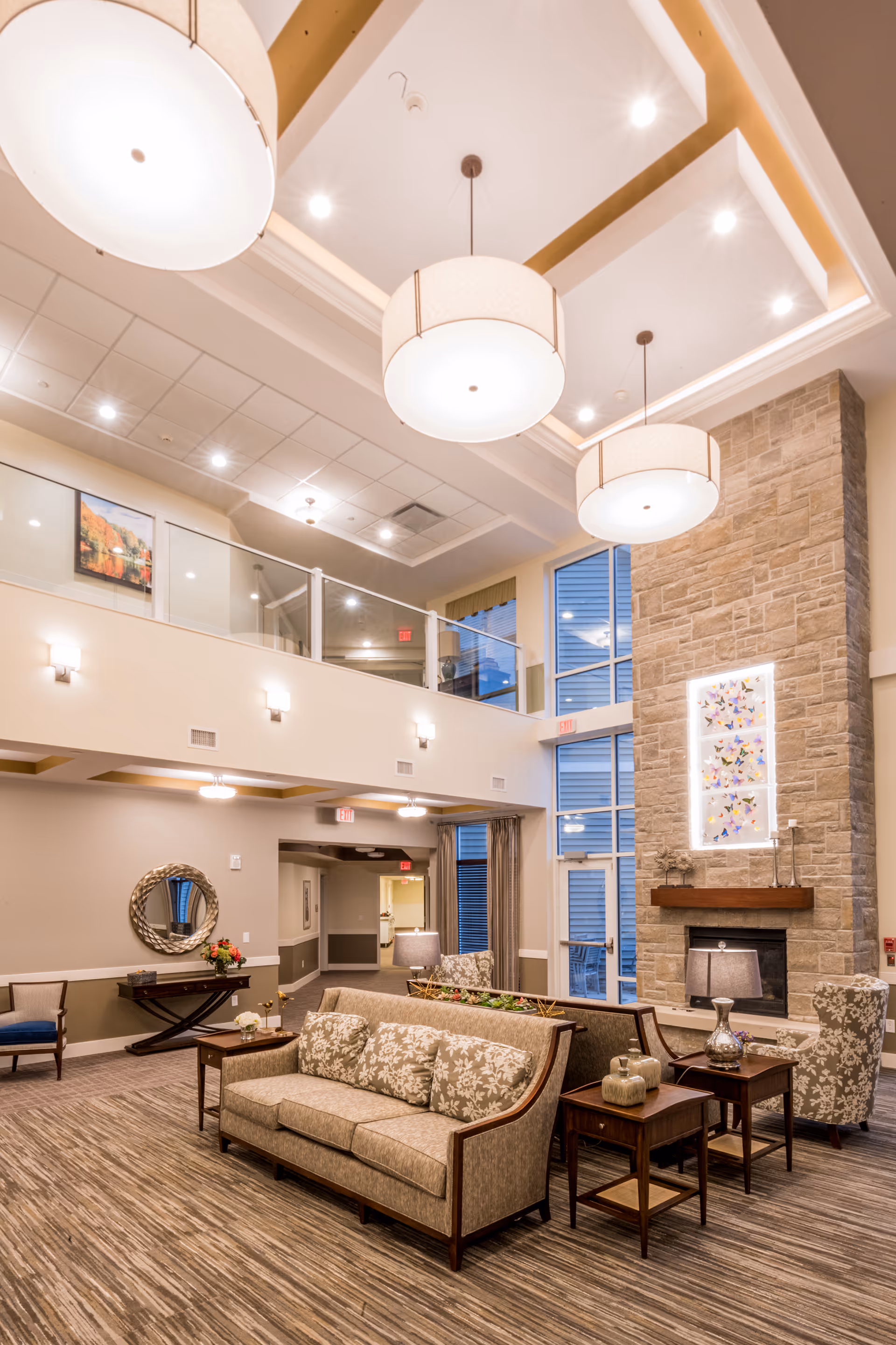 Spacious two-story senior living lounge with seating, high ceilings, pendant lights, and a stone fireplace.