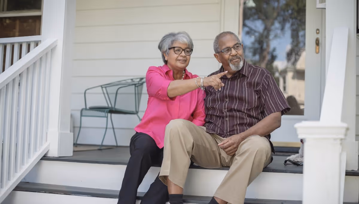 Two elderly adults sit on a porch, smiling as the woman points ahead.
