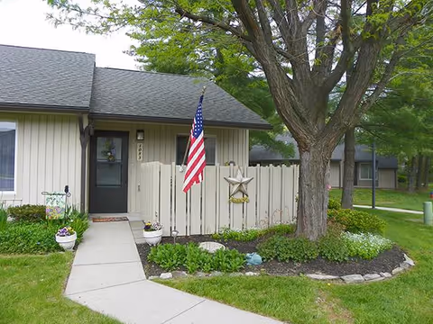 Exterior front of a ground-level residential unit with a small fenced patio, American flag, tree, and walkway.