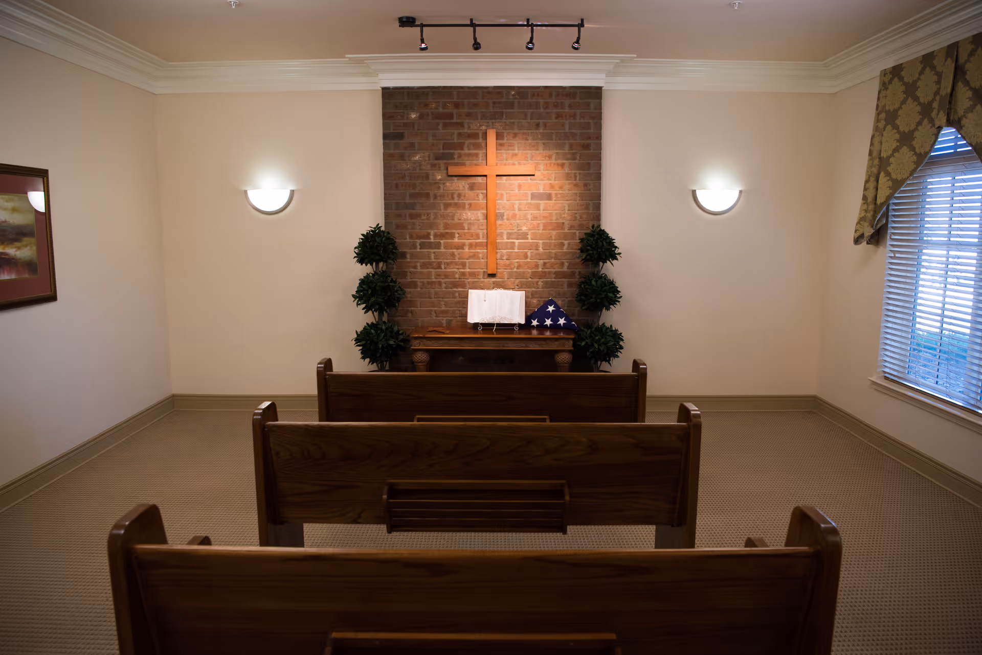 Small chapel room with three wooden pews facing a brick wall with a wooden cross mounted on it. Below the cross is a small table with a folded American flag and an open book. Two potted plants flank the table. The room has beige walls, carpeted floor, a window with patterned curtains on the right, and a framed painting on the left wall.
