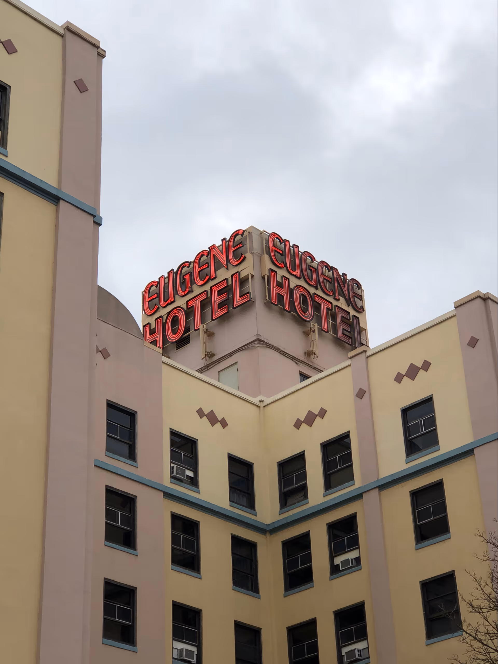 Pastel multi-story building with a red neon Eugene Hotel sign atop the front facade.