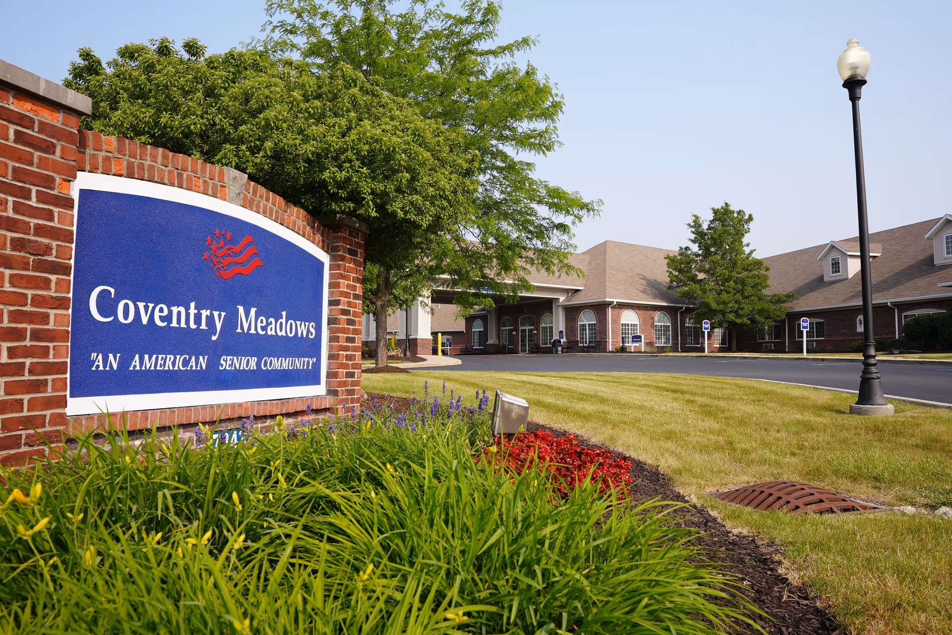 Exterior view of Coventry Meadows senior living community with a brick sign in the foreground displaying the name and slogan 'An American Senior Community'. The building is visible in the background with a driveway, green lawn, trees, and a street lamp.