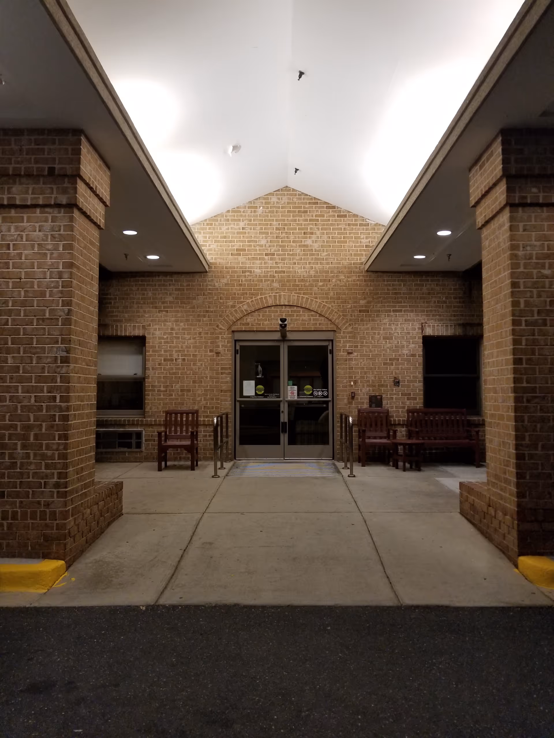Brick building entrance with double doors, covered walkway, and benches on either side.