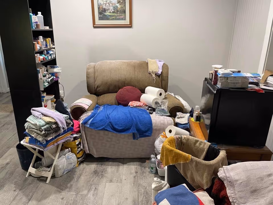 Interior living room cluttered with a sofa, shelves, blankets, and scattered supplies.