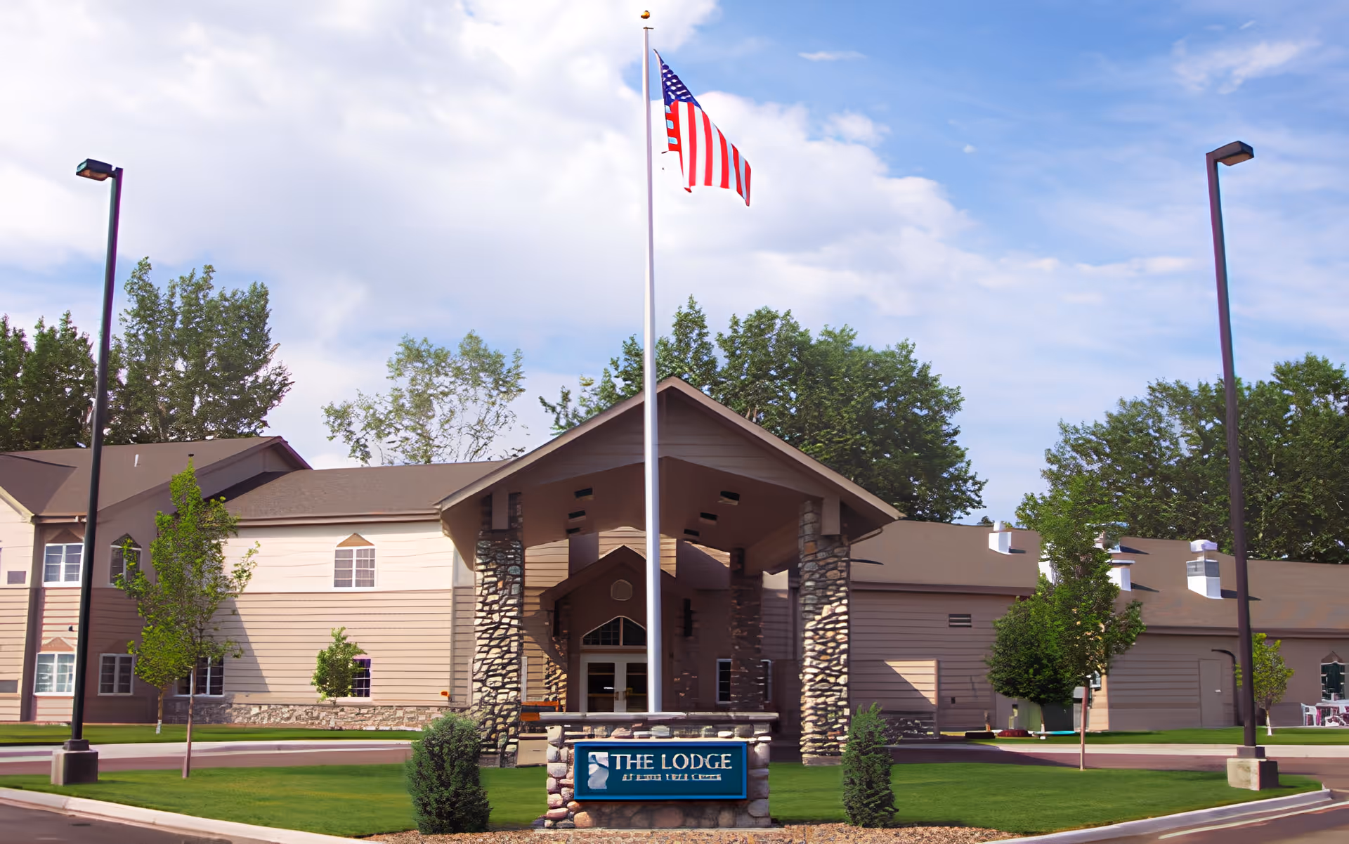 Front entrance of The Lodge senior living building with a tall flagpole flying an American flag and a stone sign on a manicured lawn.