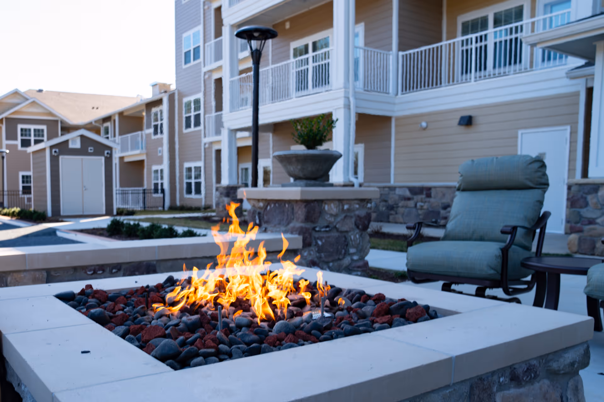 Outdoor courtyard with a lit stone fire pit, cushioned lounge chairs, and multi-story apartment balconies in the background.