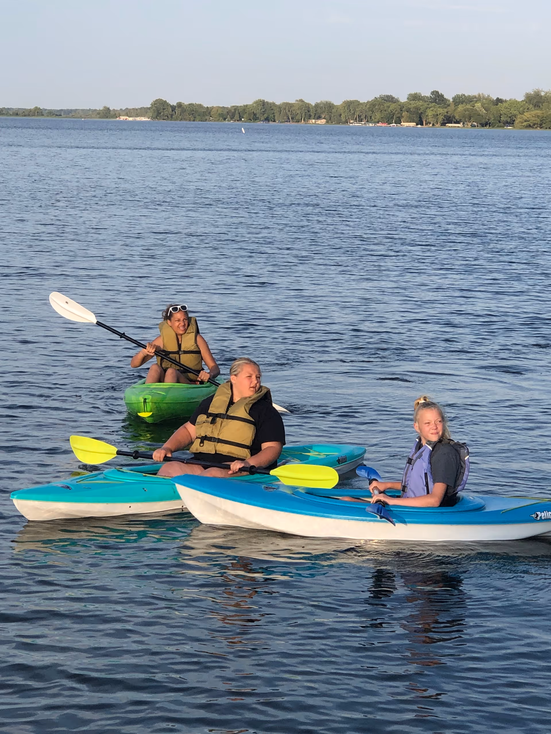 Three people in kayaks wearing life jackets paddle on a calm lake.