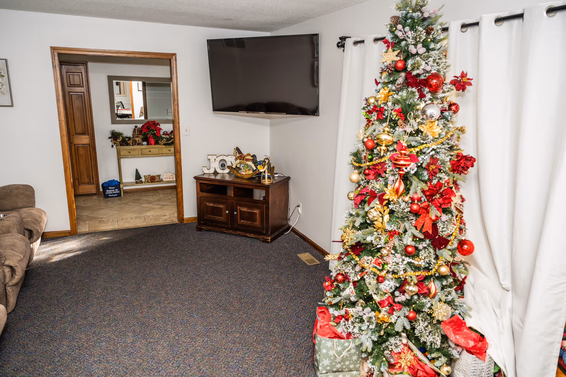 A living room decorated for Christmas with a tall Christmas tree adorned with red, gold, and white ornaments and surrounded by wrapped presents. There is a brown couch on the left, a wooden TV stand with holiday decorations including a 'JOY' sign, and a flat-screen TV mounted on the wall. In the background, there is a hallway with a console table decorated with holiday items and a mirror above it. White curtains cover the window on the right side.