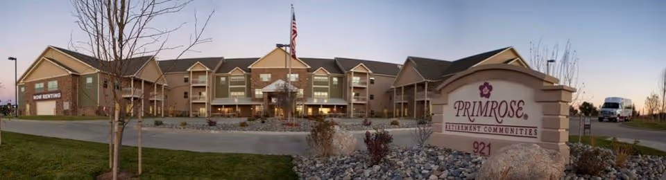 Front exterior of the Primrose Retirement Community of Gillette with its entrance, flagpole, and a landscaped sign in the foreground.