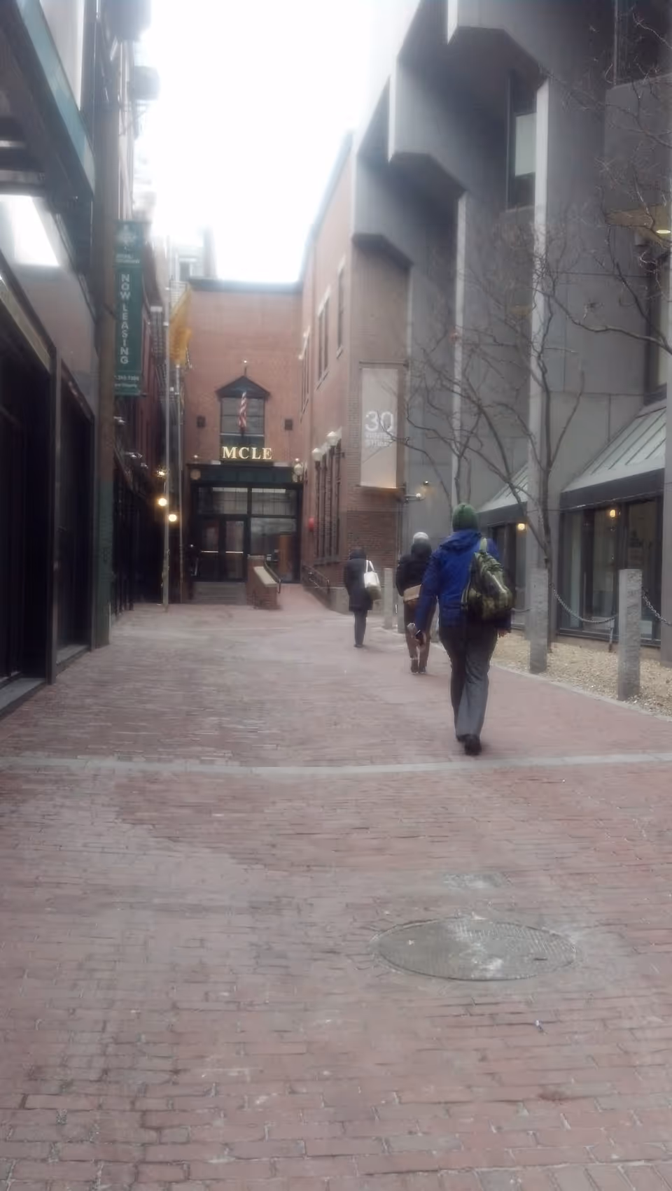 Outdoor brick-paved walkway between tall buildings with people walking toward an entrance.