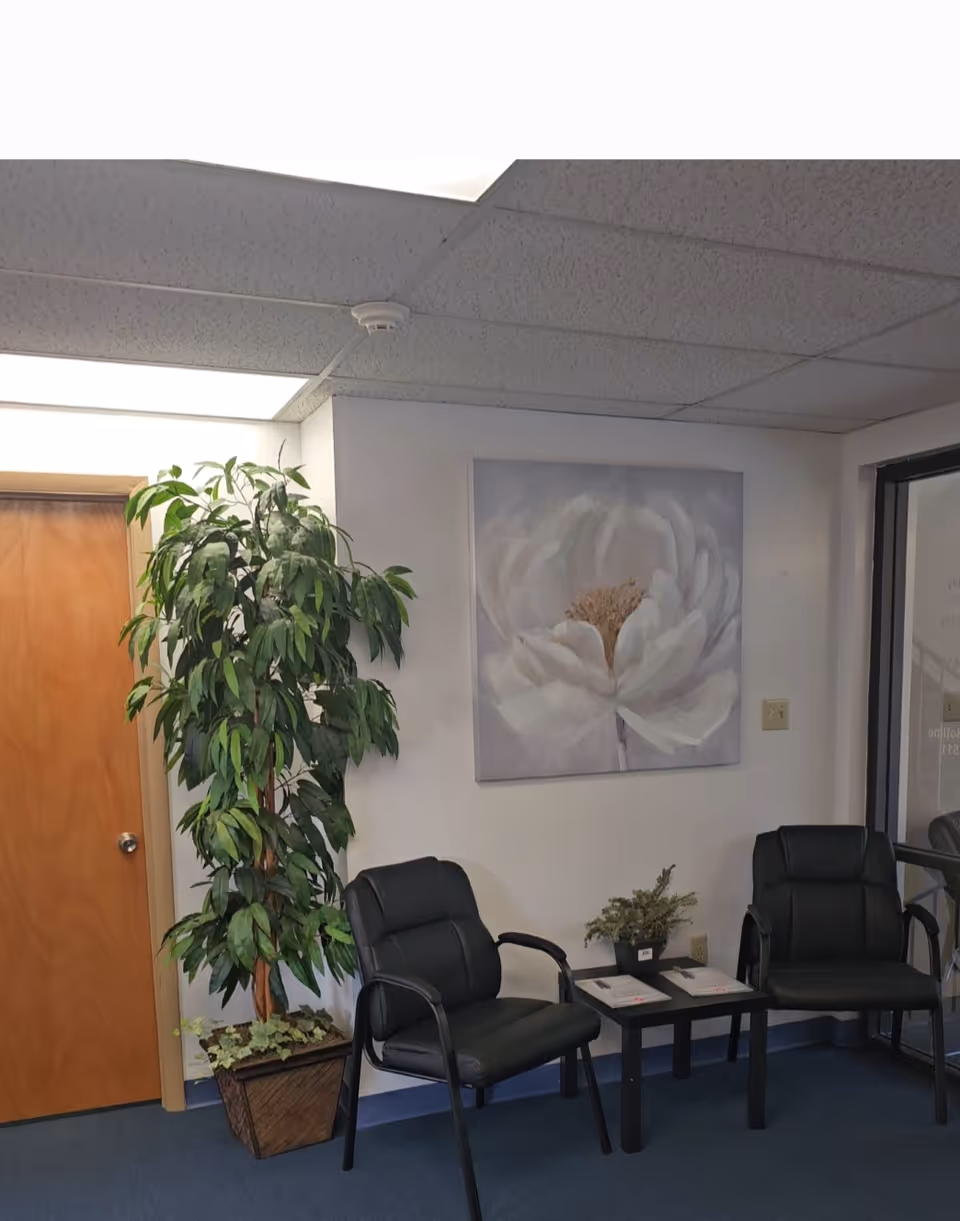 Office waiting area with two black chairs, small table, plant, and floral artwork.