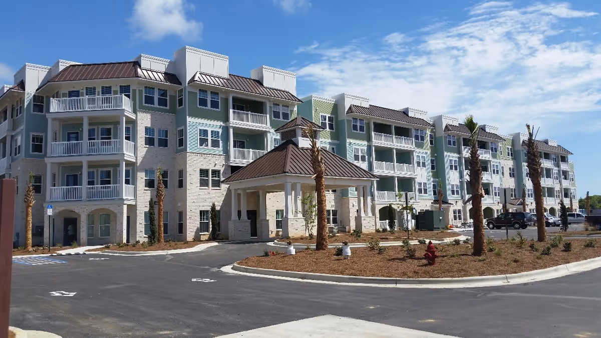 Exterior view of a multi-story senior living facility with balconies, a covered entrance, palm trees, and a parking area under a partly cloudy sky.