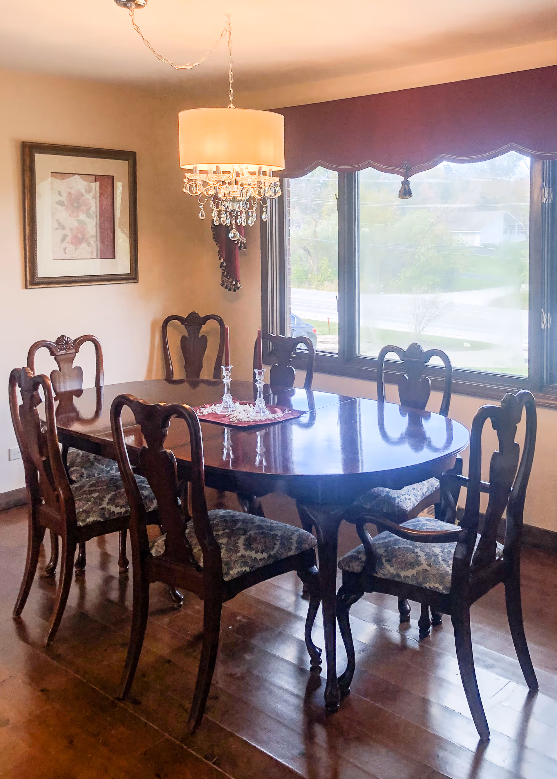 A dining room with a polished wooden oval table surrounded by six wooden chairs with patterned cushions. A chandelier with crystal decorations hangs above the table. There is a large window with a maroon valance letting in natural light, and a framed floral picture on the wall.
