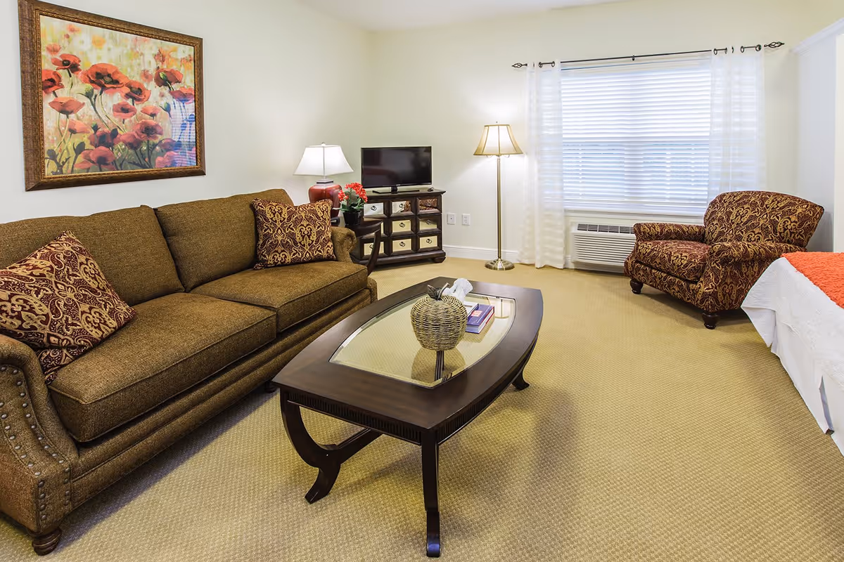 A cozy senior living room featuring a brown upholstered sofa with patterned cushions, a matching armchair, a wooden coffee table with a glass top and decorative items, a small TV on a wooden cabinet, a floor lamp, a table lamp, and a large window with white blinds and sheer curtains. A colorful floral painting hangs on the wall above the sofa.