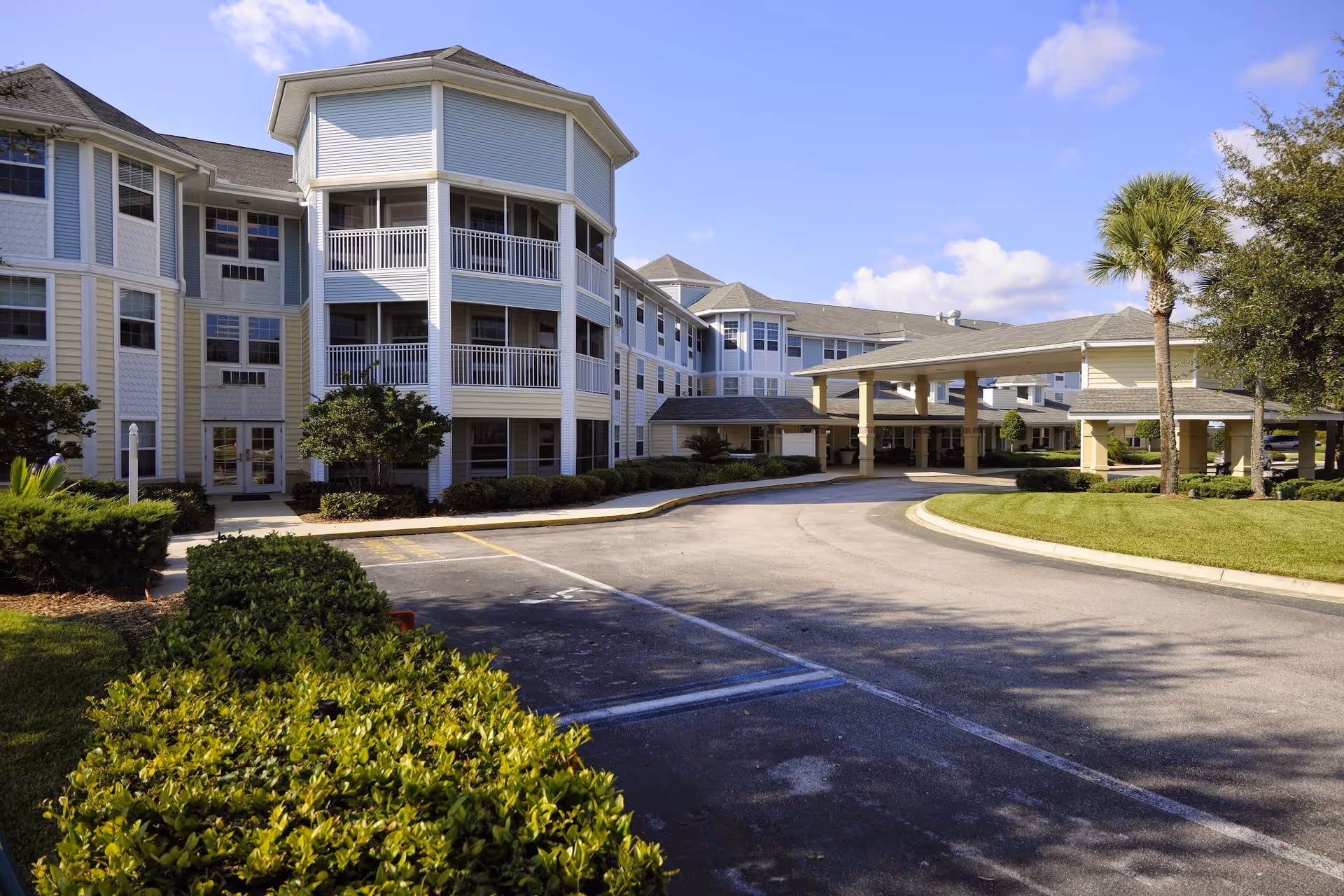 Front exterior of a multi-story senior living building with balconies, a covered porte-cochère, circular driveway, and landscaped grounds.