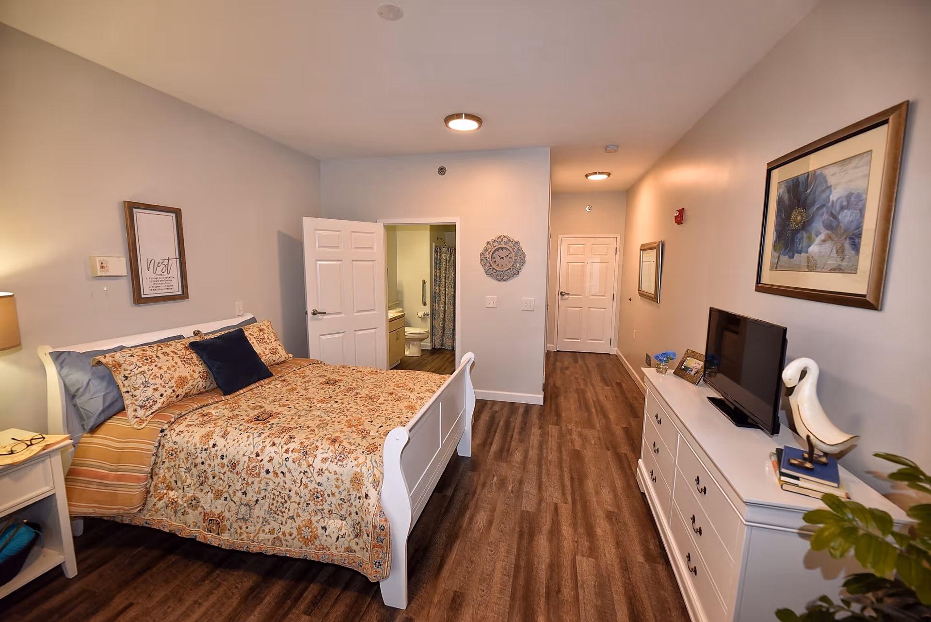 Bright bedroom featuring a white bed, dresser, TV, and a hallway to a bathroom.