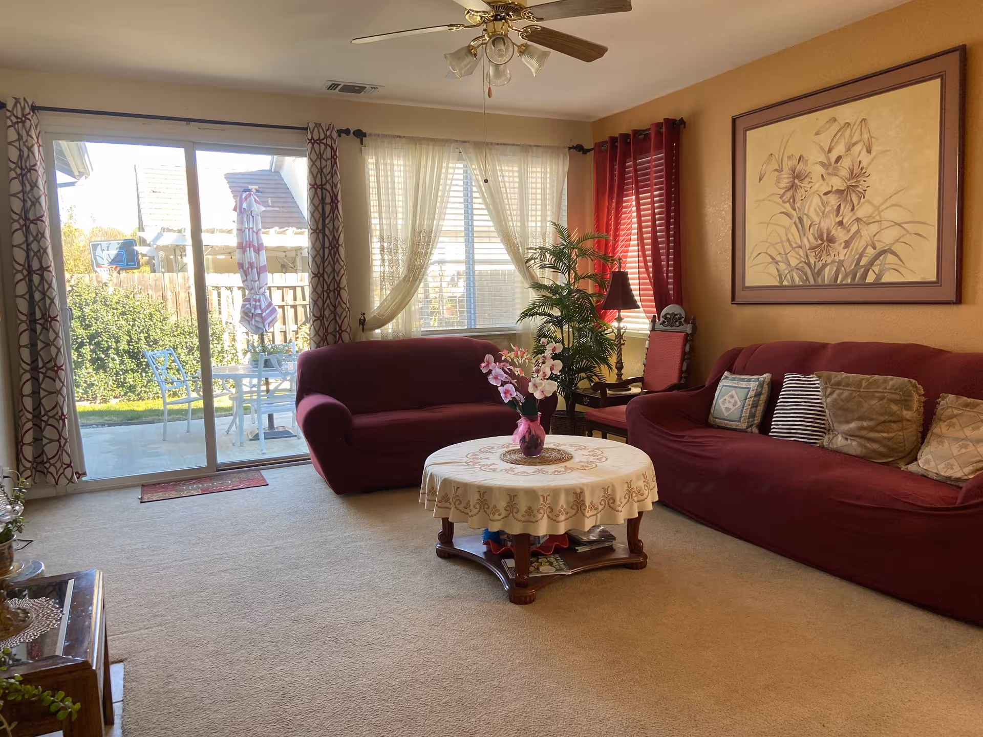 A cozy living room with beige carpet and walls painted in a warm tone. The room features two maroon sofas with assorted cushions, a round wooden coffee table covered with a lace tablecloth and a vase of flowers on top. There is a large window with sheer curtains and red drapes, and a sliding glass door leading to an outdoor patio with a table, chairs, and an umbrella. A ceiling fan with lights is mounted on the ceiling, and a framed floral painting hangs on the wall above one sofa.