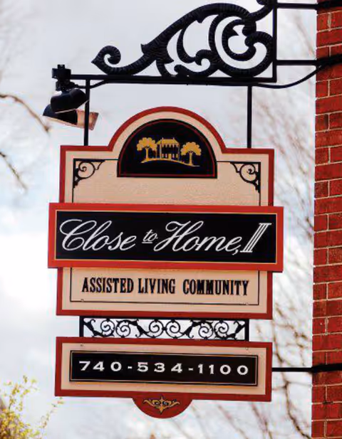 Decorative hanging sign reading "Close to Home II" Assisted Living Community with a phone number mounted on a brick exterior.