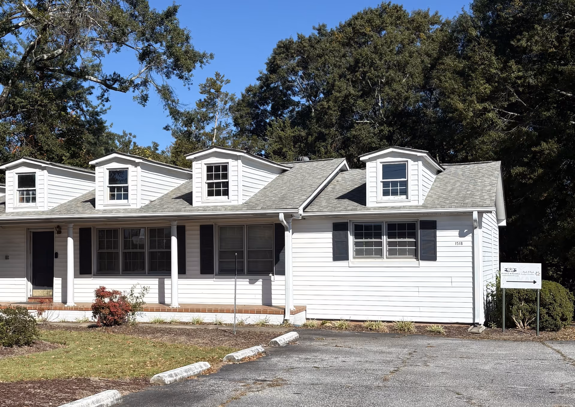 White single-story building with dormer windows, black shutters, and a facility sign by the driveway.