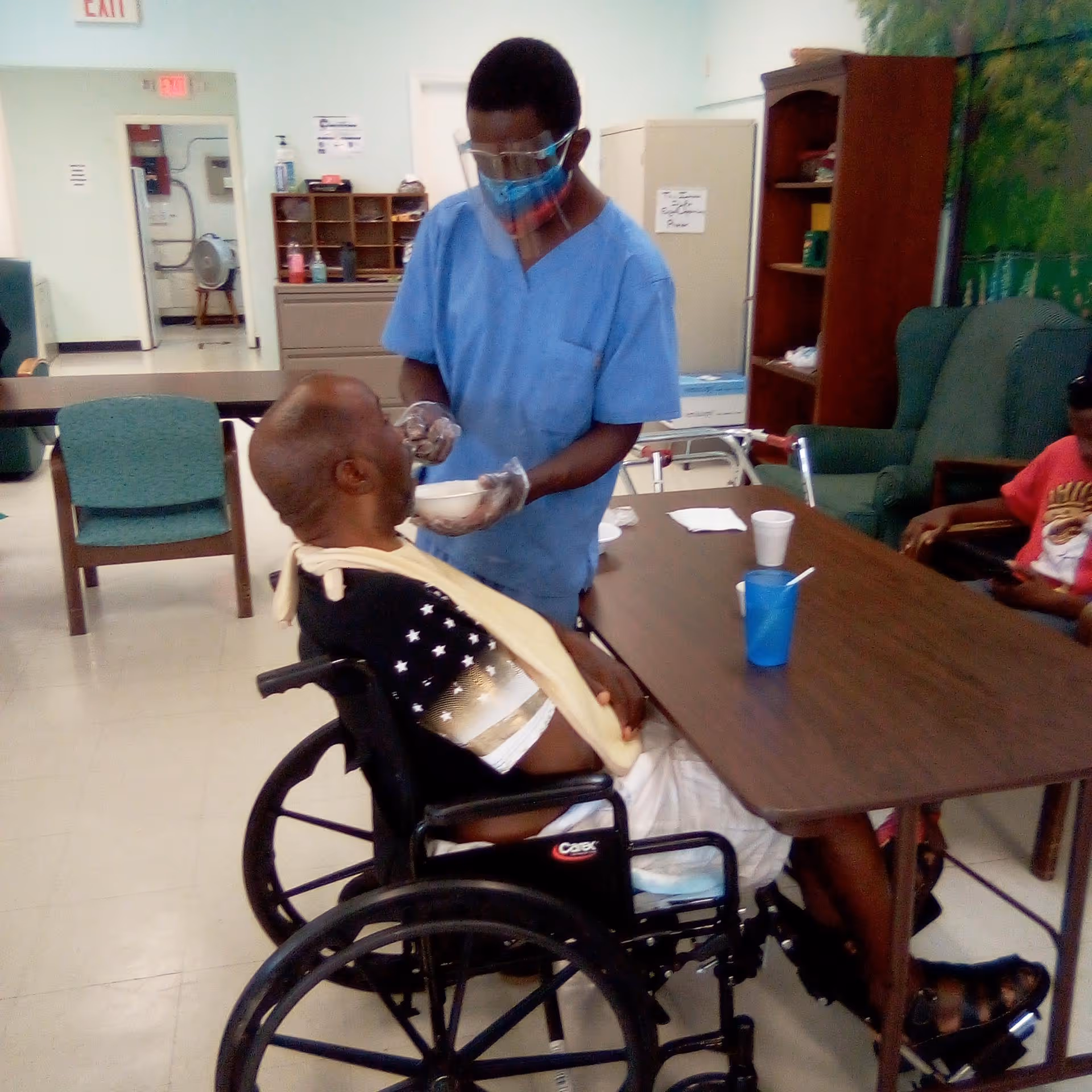 Caregiver feeds a man in a wheelchair at a dining table.