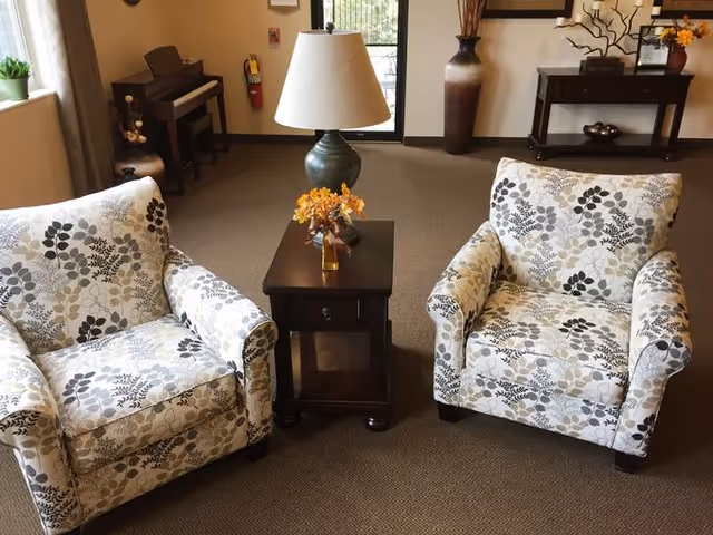 Two patterned armchairs with floral designs are positioned on either side of a small wooden side table with a lamp and a small vase of orange flowers. In the background, there is a piano, a fire extinguisher mounted on the wall, a large vase with decorative branches, and a console table with decorative items. The room has carpeted flooring and a door leading outside.