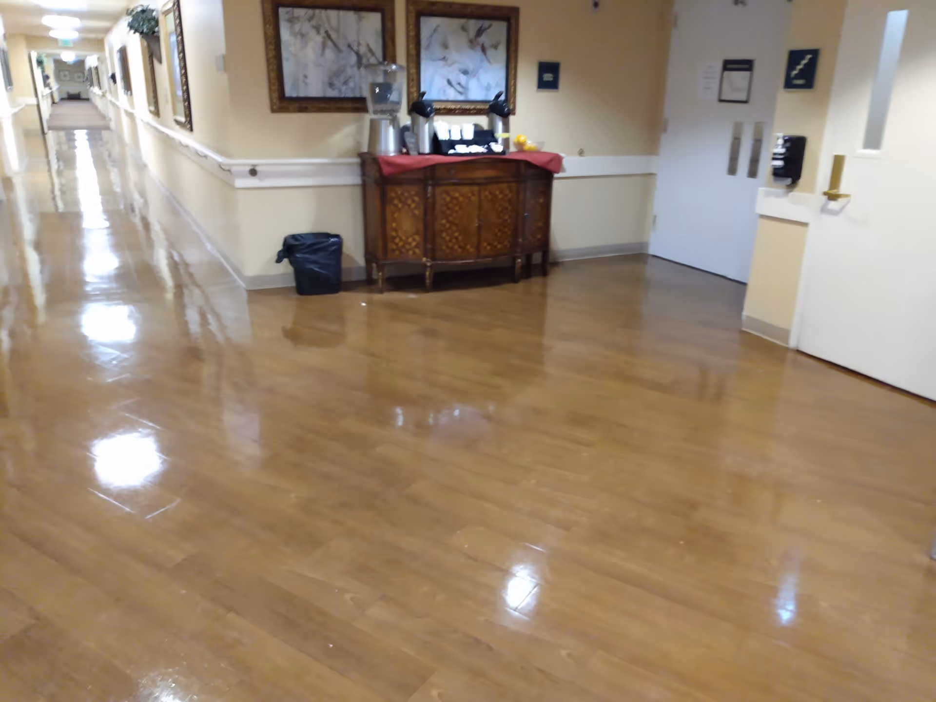 Long polished interior hallway with a wooden console holding a coffee station, framed artwork, and a trash can.