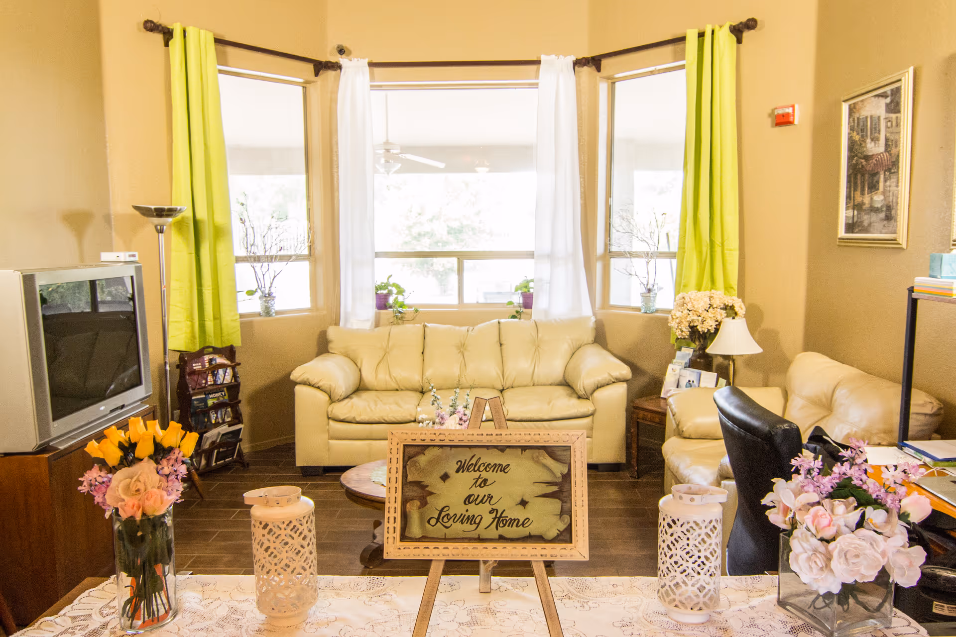 A cozy living room with beige leather sofas, a wooden coffee table, and a TV on a wooden stand. The room has three windows with green and white curtains, and several flower arrangements and decorative items are placed around the room. A framed sign on an easel reads 'Welcome to our Loving Home.'