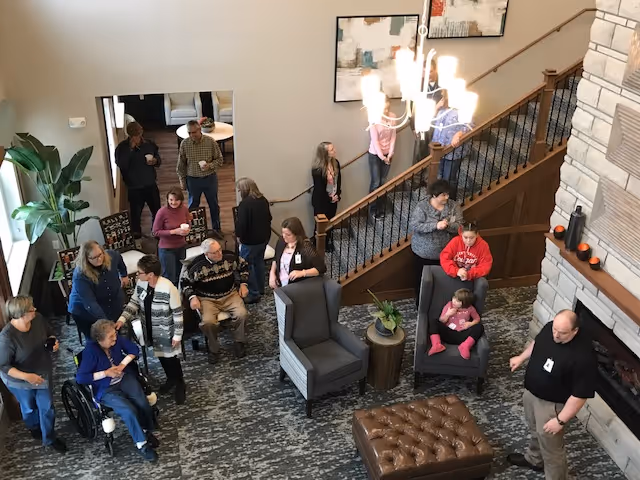 A group of people socializing in a spacious living room area with carpeted floors, two gray armchairs, a brown tufted ottoman, and a stone fireplace. Some people are seated while others stand or walk near a staircase with a wooden handrail. The room has modern decor including abstract paintings on the wall and a large plant near a window.