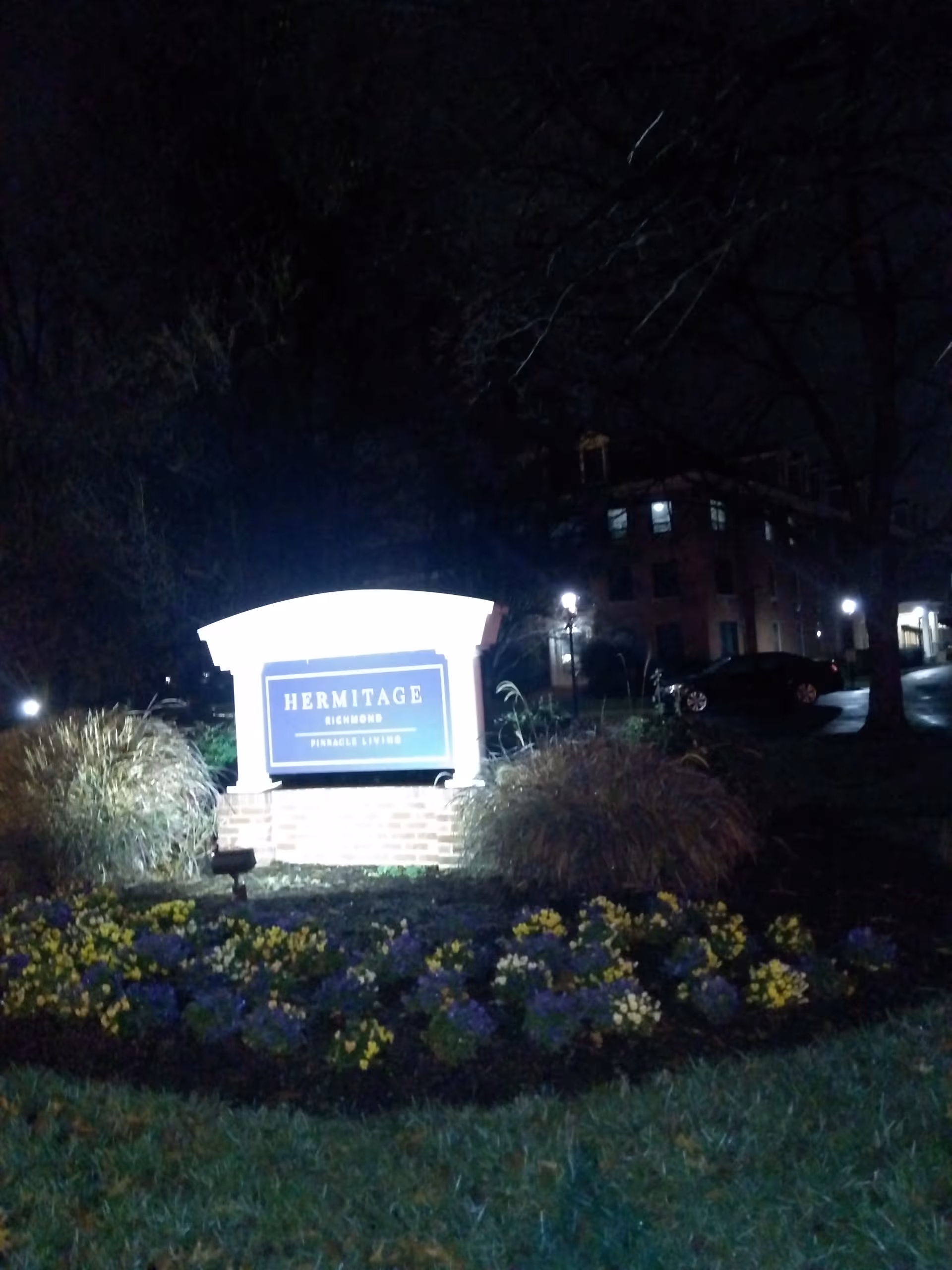 Nighttime exterior view of Hermitage Richmond sign with flowers and building in background.