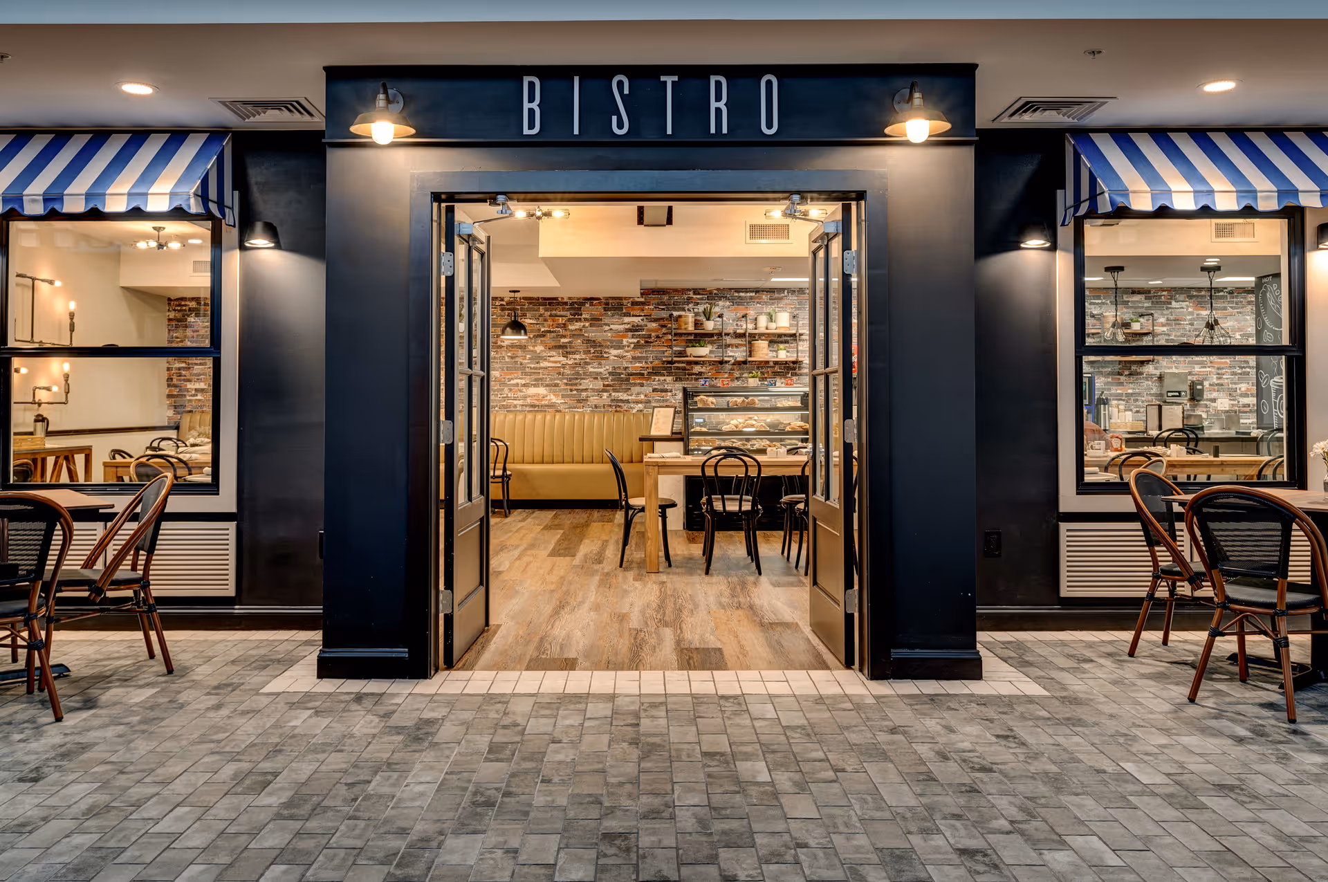 Entrance to a bistro with open double doors revealing a cozy interior featuring a brick accent wall, wooden tables, black chairs, and a display case with baked goods. The exterior has a tiled floor and blue and white striped awnings over the windows on either side of the entrance.