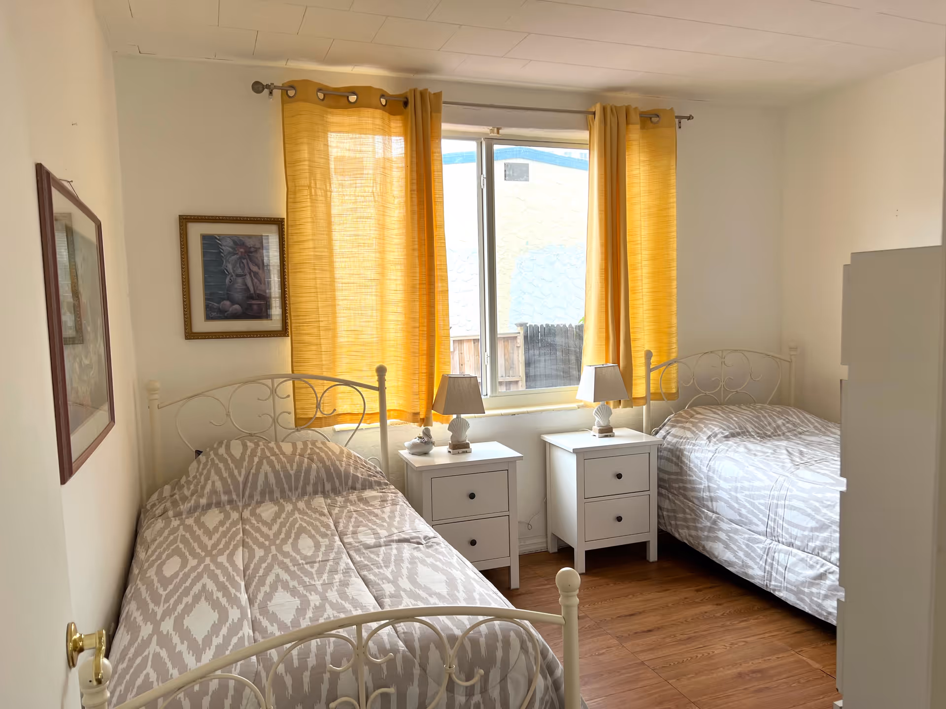 Two twin beds with white metal frames and yellow curtains in a bright bedroom.