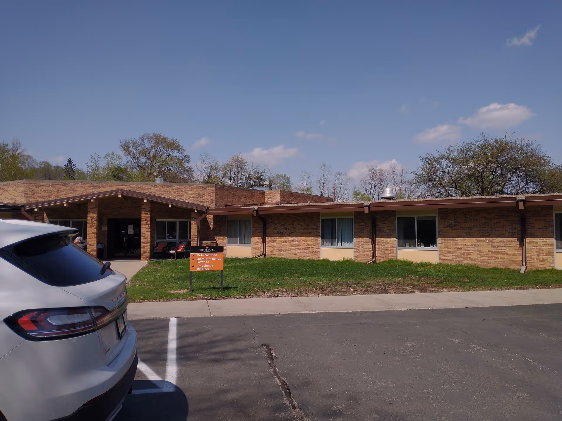 Brick senior living facility exterior with parked car and front entrance on a sunny day.