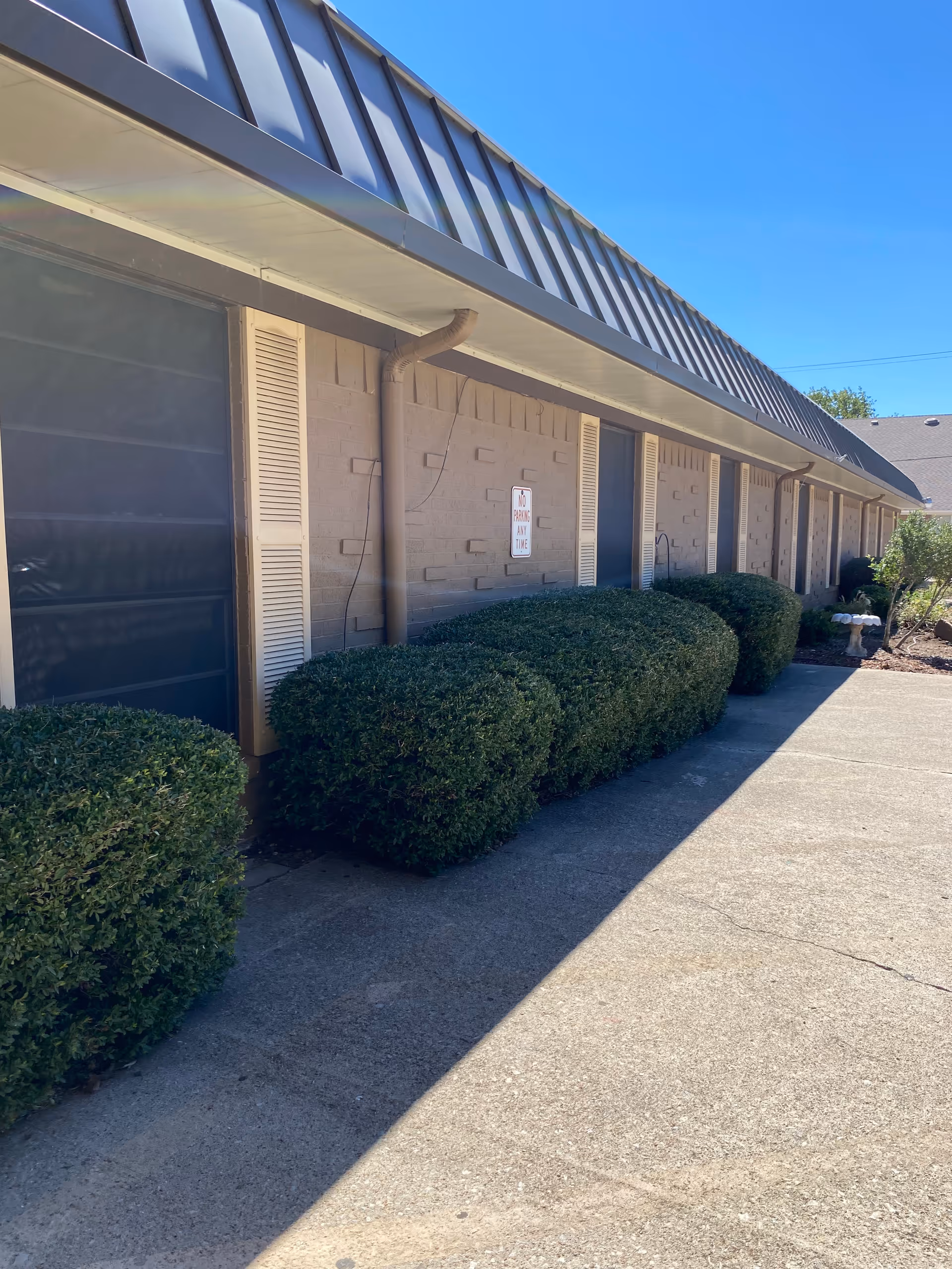 Exterior of a Resthome building with a sidewalk, hedges, and vent pipes along the wall.