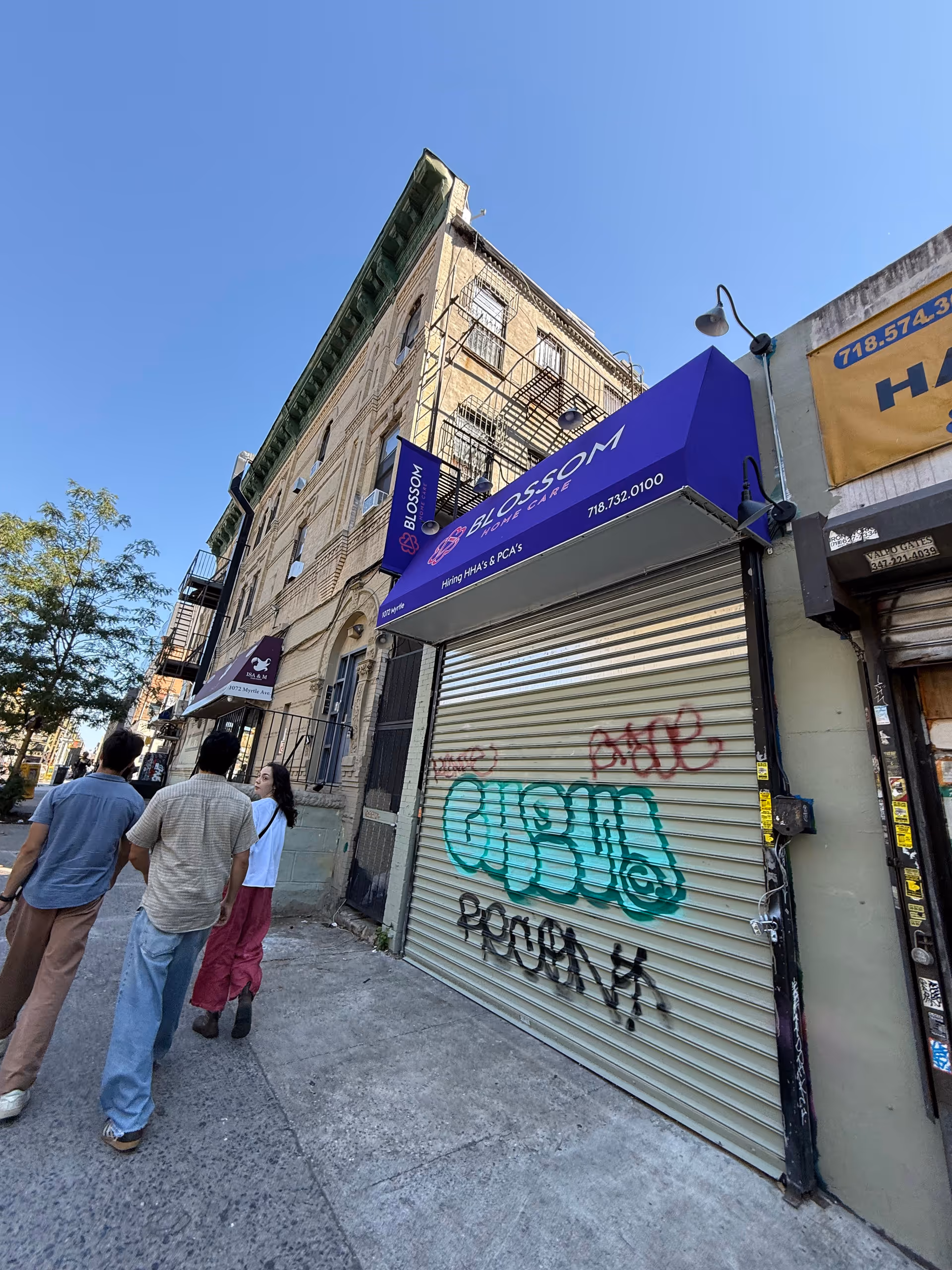 Three people stand outside a Blossom Home Care storefront on a Brooklyn street.