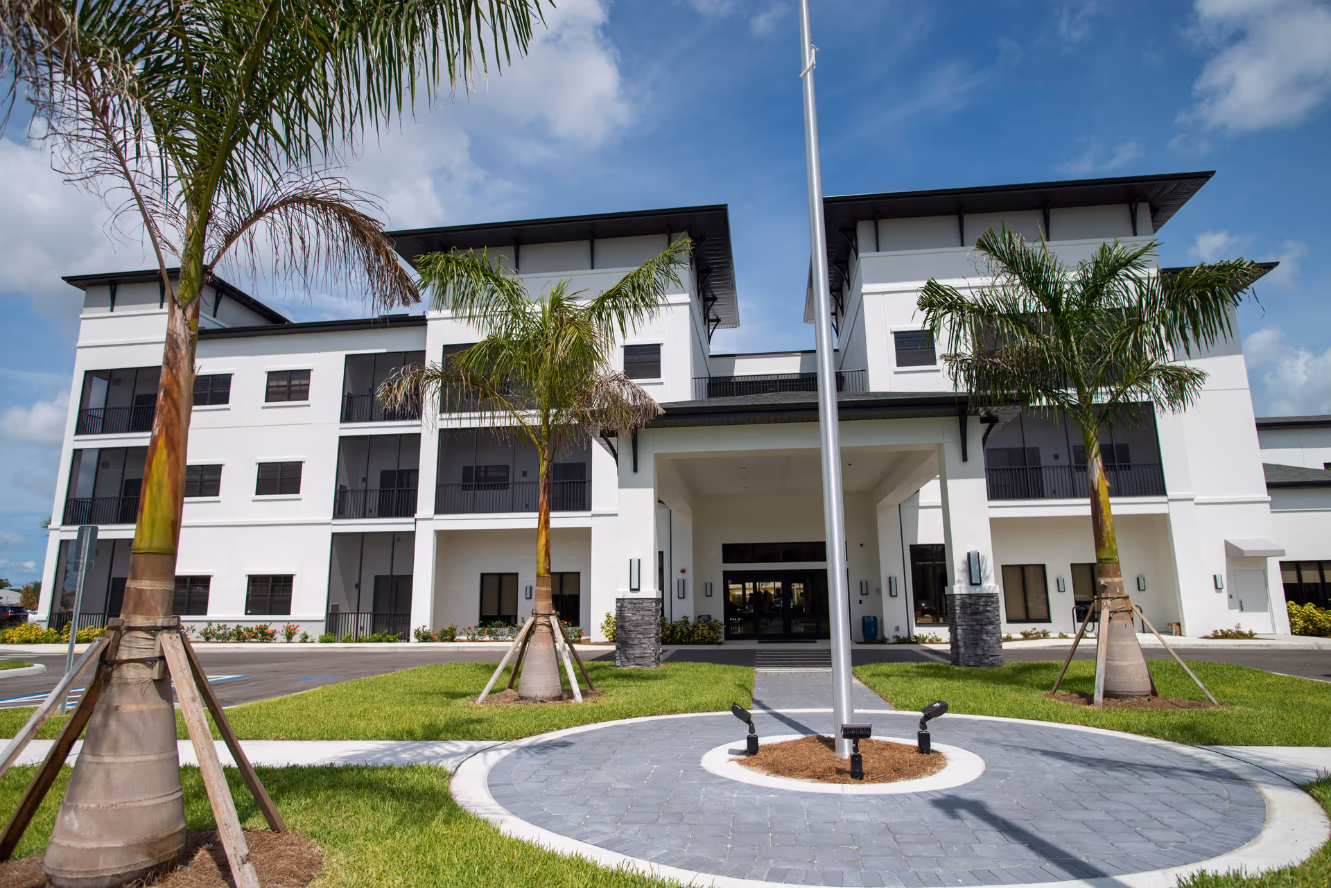 Front exterior view of a modern three-story white building with black window frames and balconies, surrounded by palm trees and a circular driveway with a flagpole in the center under a partly cloudy blue sky.
