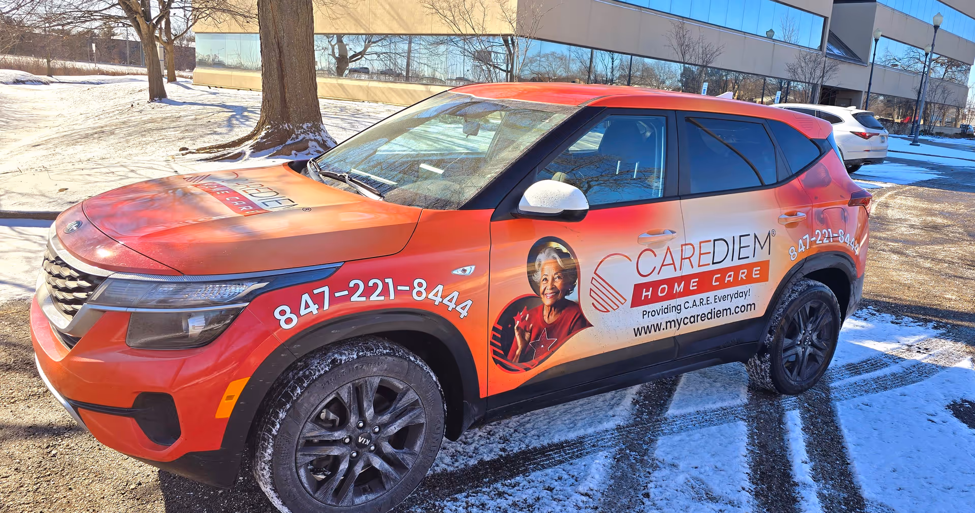 A red CareDiem Home Care SUV parked in snowy lot with a modern building in background.