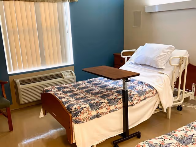 A small room with a hospital-style bed covered with a floral patterned blanket and white sheets. There is a wooden bedside table with drawers, a movable overbed table, a window with vertical blinds, an air conditioning unit below the window, and a wooden chair partially visible on the left side.